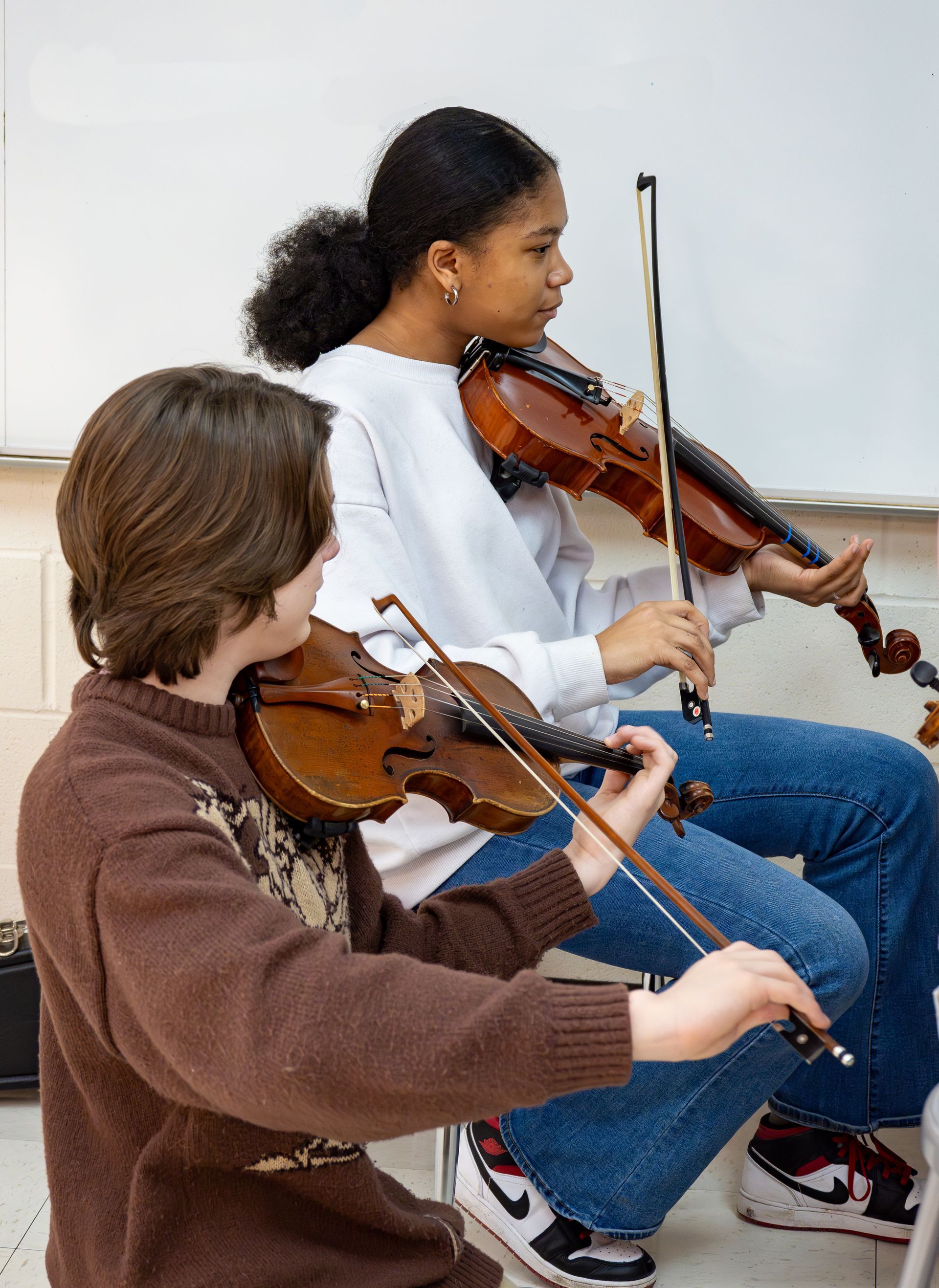Two people in a classroom practicing the violin, focused on their instruments while seated.