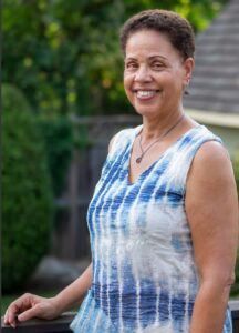 A smiling person wearing a blue and white tie-dye sleeveless top, standing outdoors with a blurred green background.