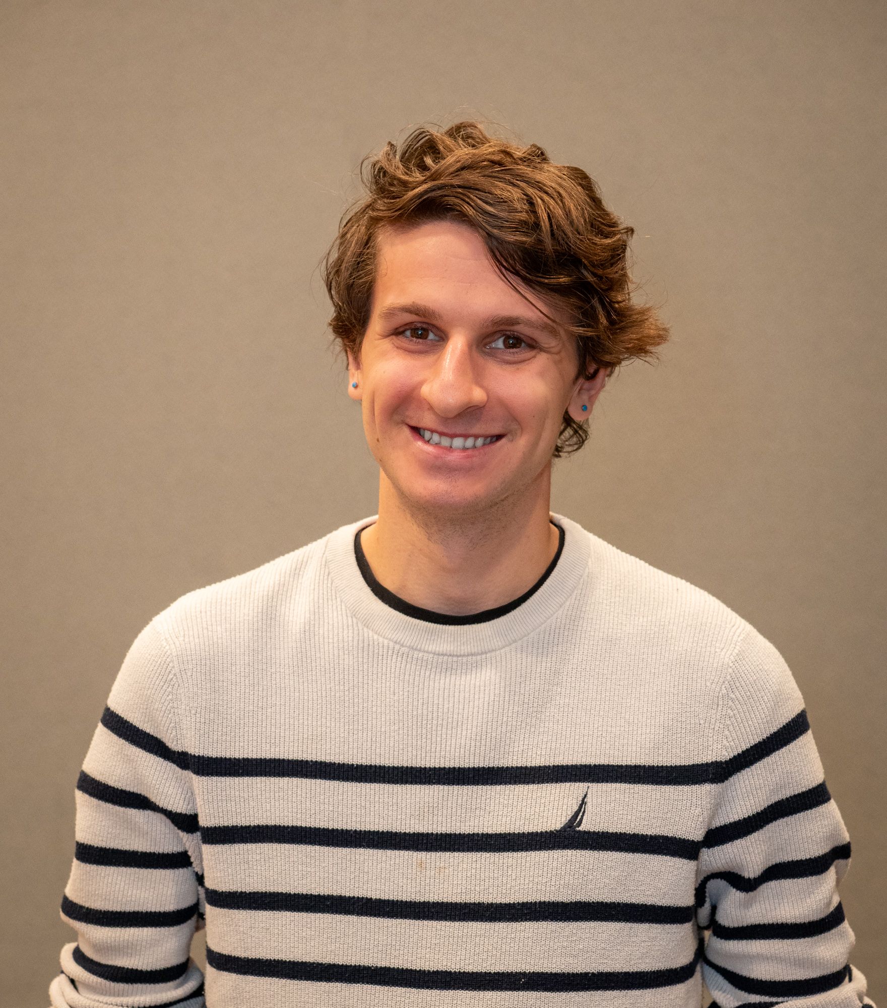 A smiling person with wavy brown hair wearing a white and dark blue striped sweater against a plain gray background.
