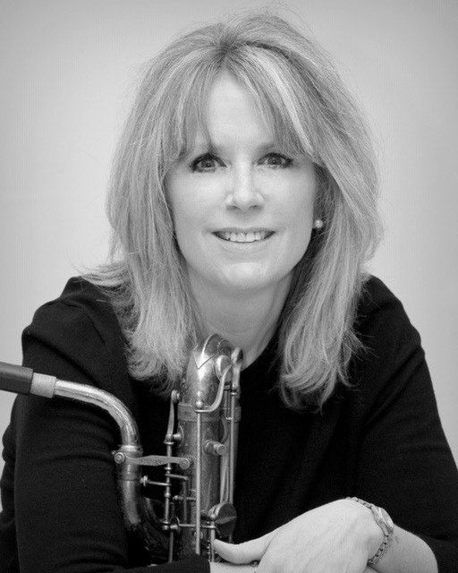 A person with shoulder-length hair poses in a black top, holding a saxophone. Black and white, portrait-style.