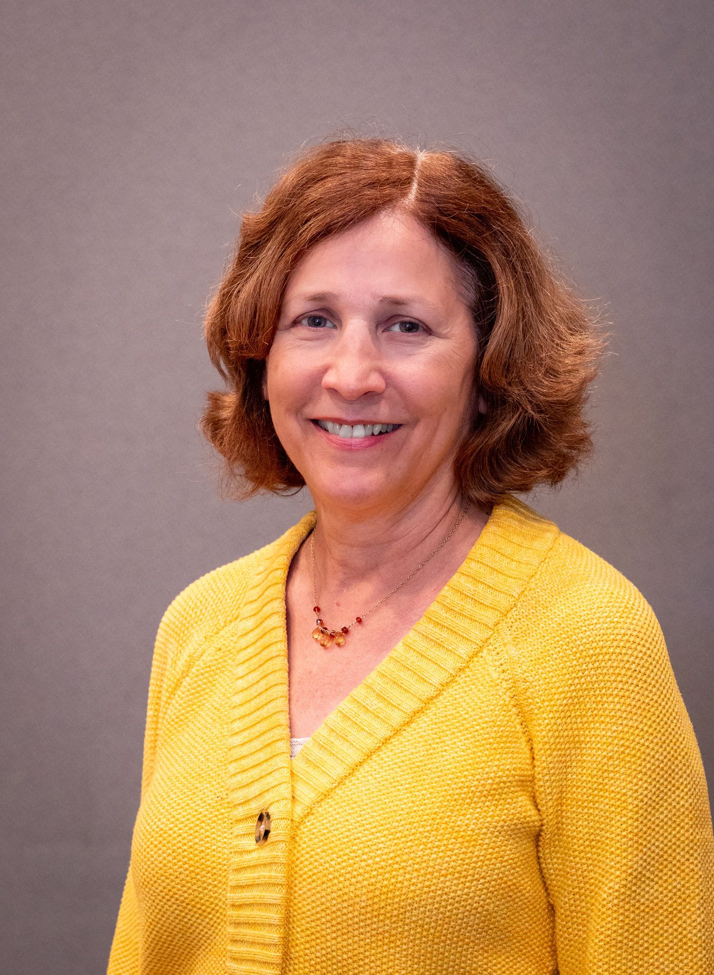 A person with shoulder-length wavy brown hair smiling, wearing a yellow knitted cardigan and a simple necklace.