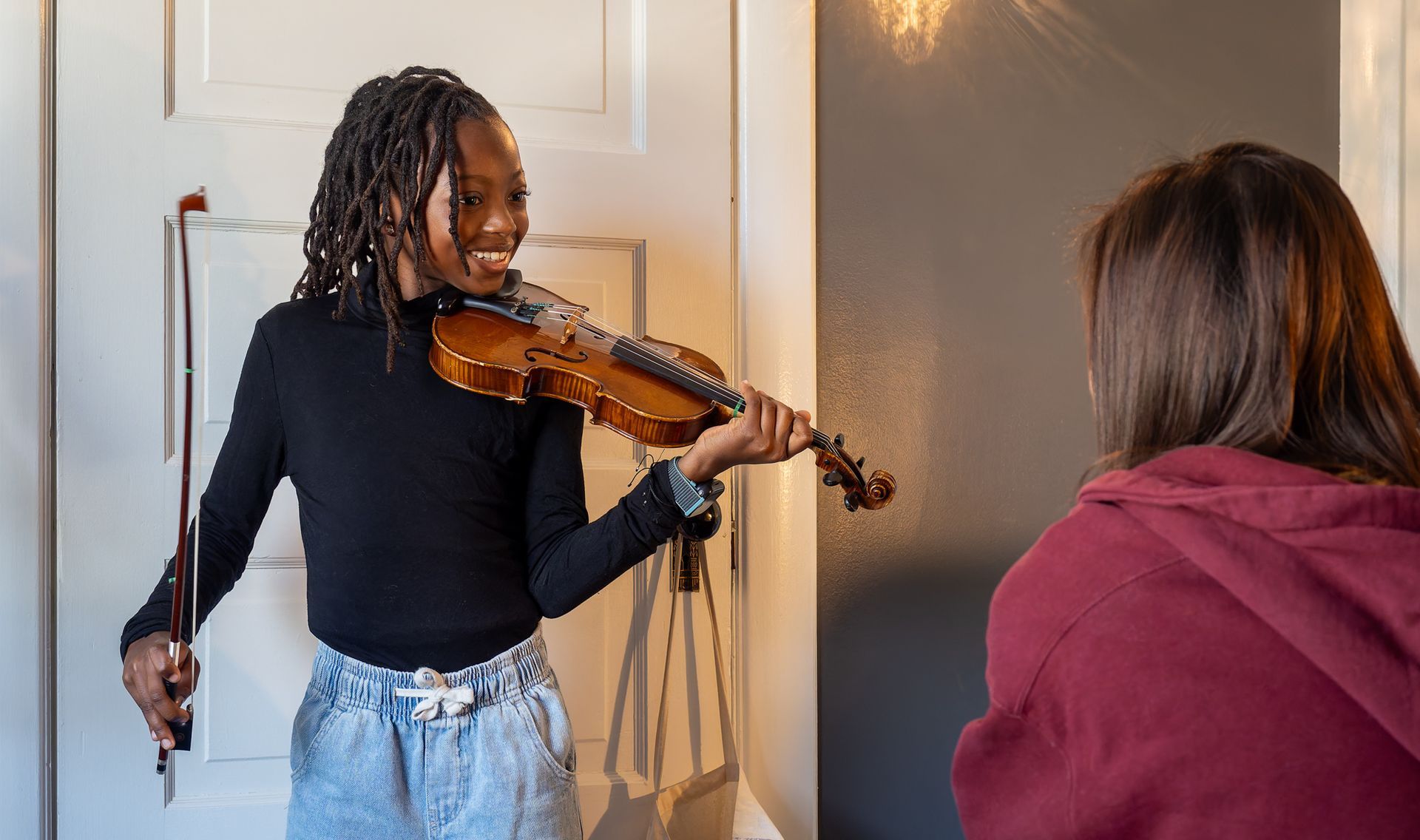 A person smiling while holding a violin and bow, facing another individual in a room with a white door.
