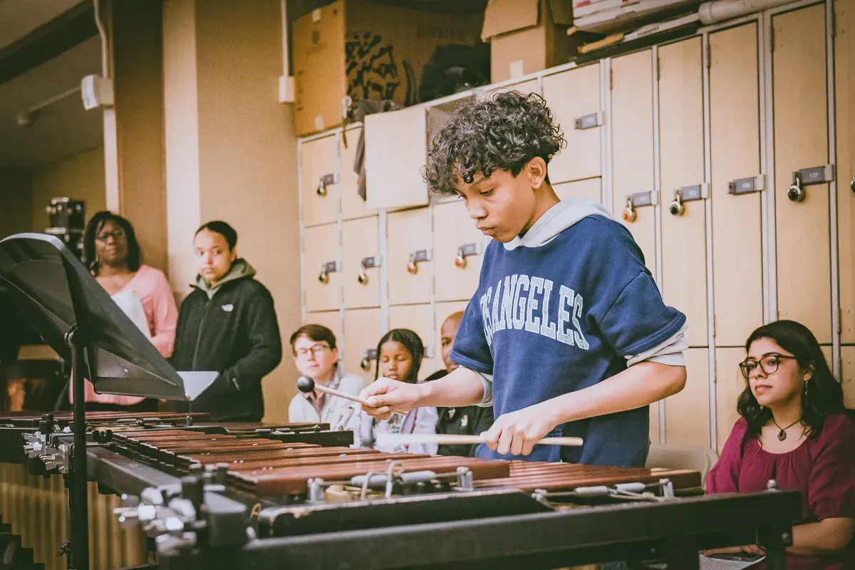 A student plays a marimba in a classroom, with peers and an instructor watching nearby in front of school lockers.