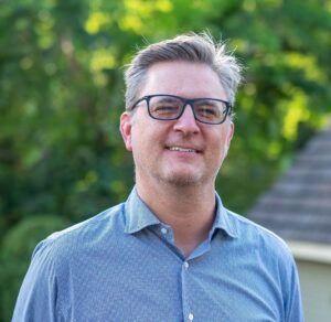A person wearing glasses and a blue button-down shirt smiling outdoors with a blurred green background.