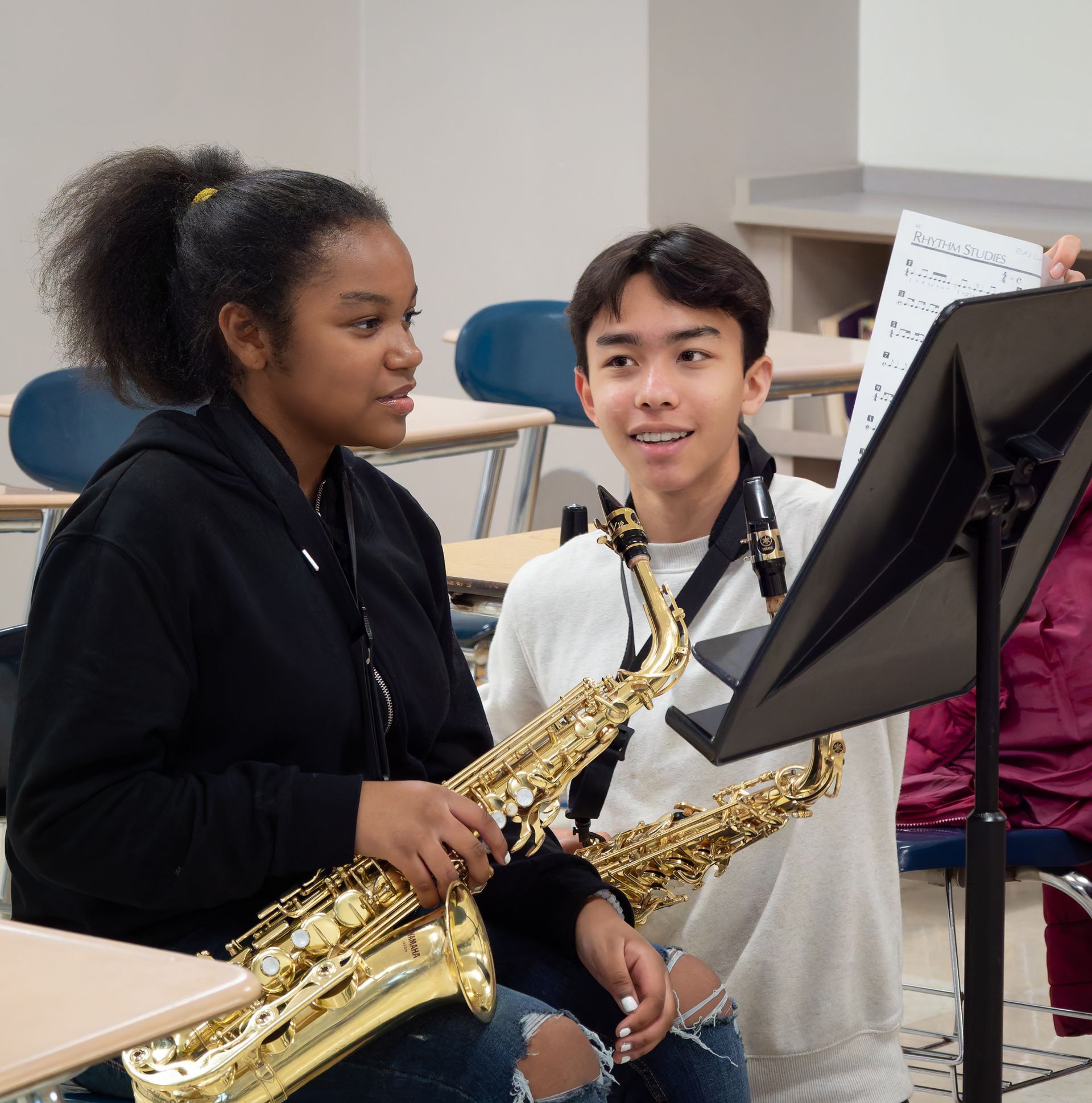 Two students holding saxophones in a classroom, looking together at sheet music on a music stand.