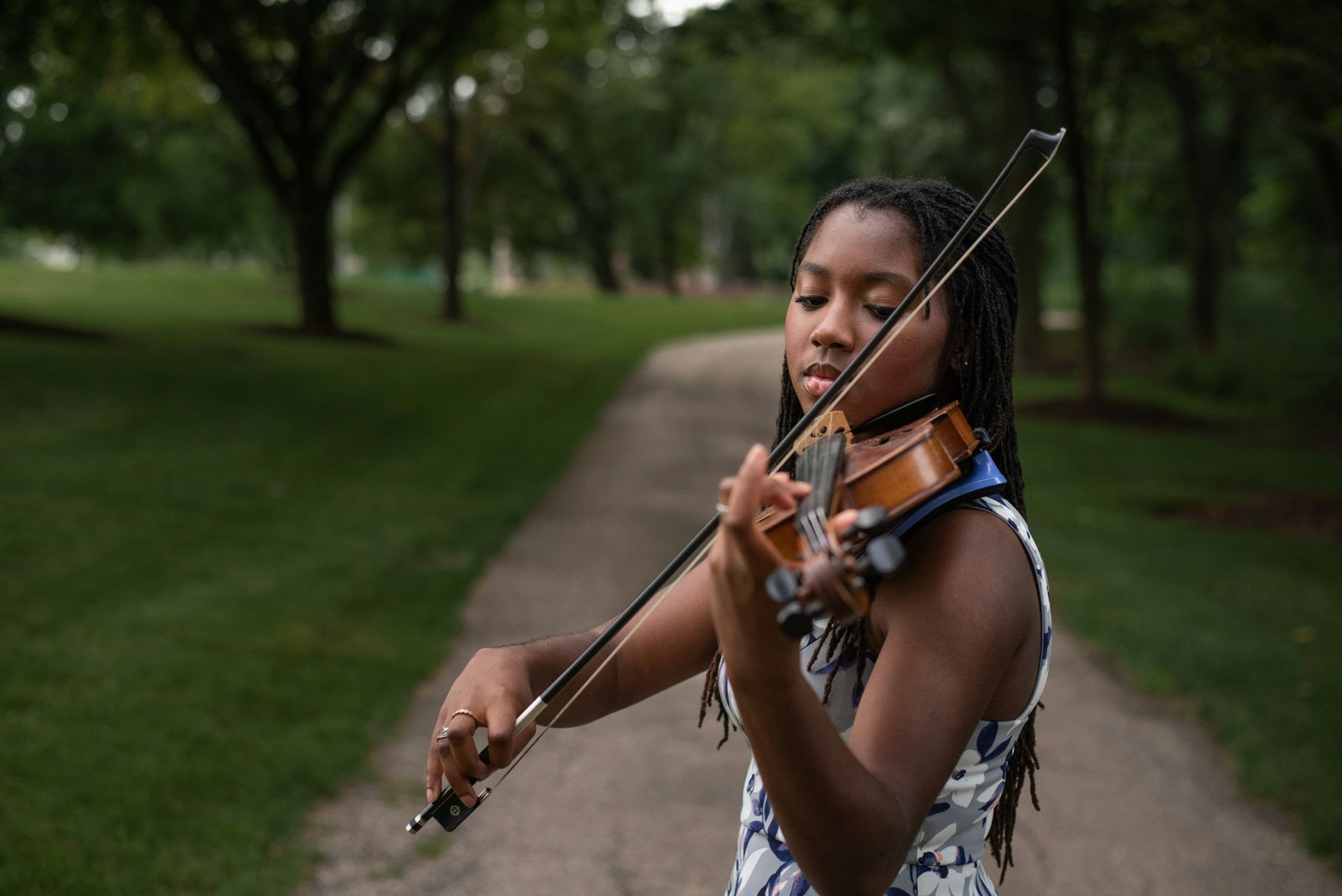 A person with braided hair plays the violin on a path in a park with green trees in the background.