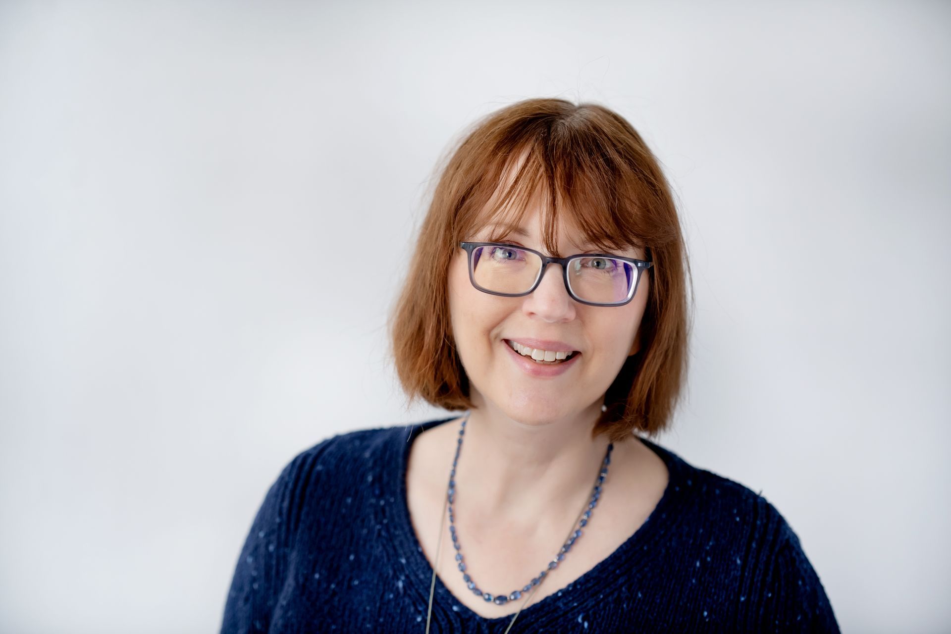A person with shoulder-length auburn hair and glasses smiling against a white background, wearing a dark blue sweater.