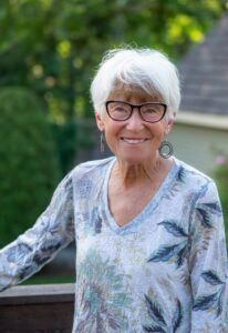 A person with short white hair and glasses smiles while wearing a long-sleeved shirt with a blue and white leafy pattern.