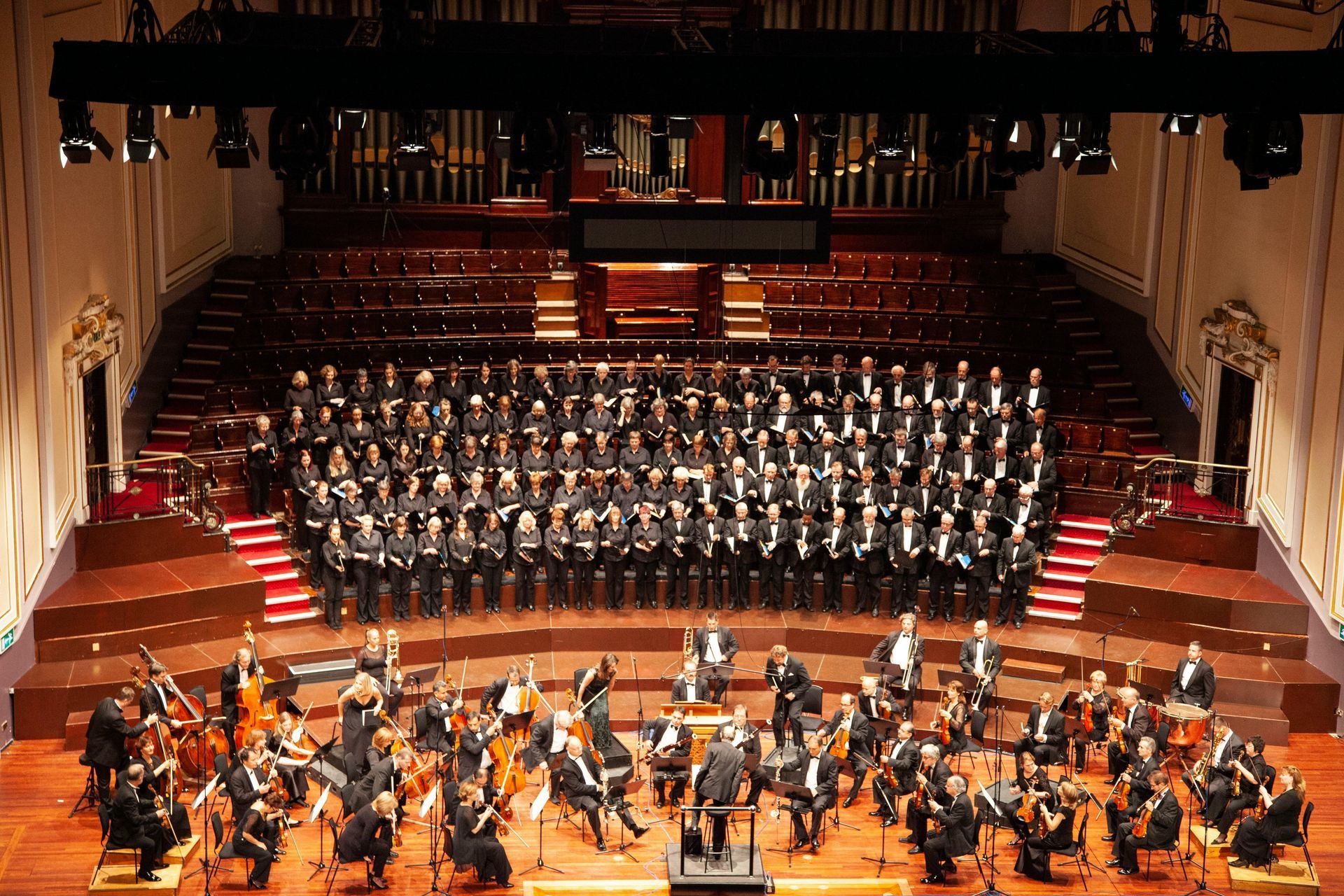 A symphony orchestra and large choir perform on a tiered wooden stage in a concert hall under theatrical spotlights.