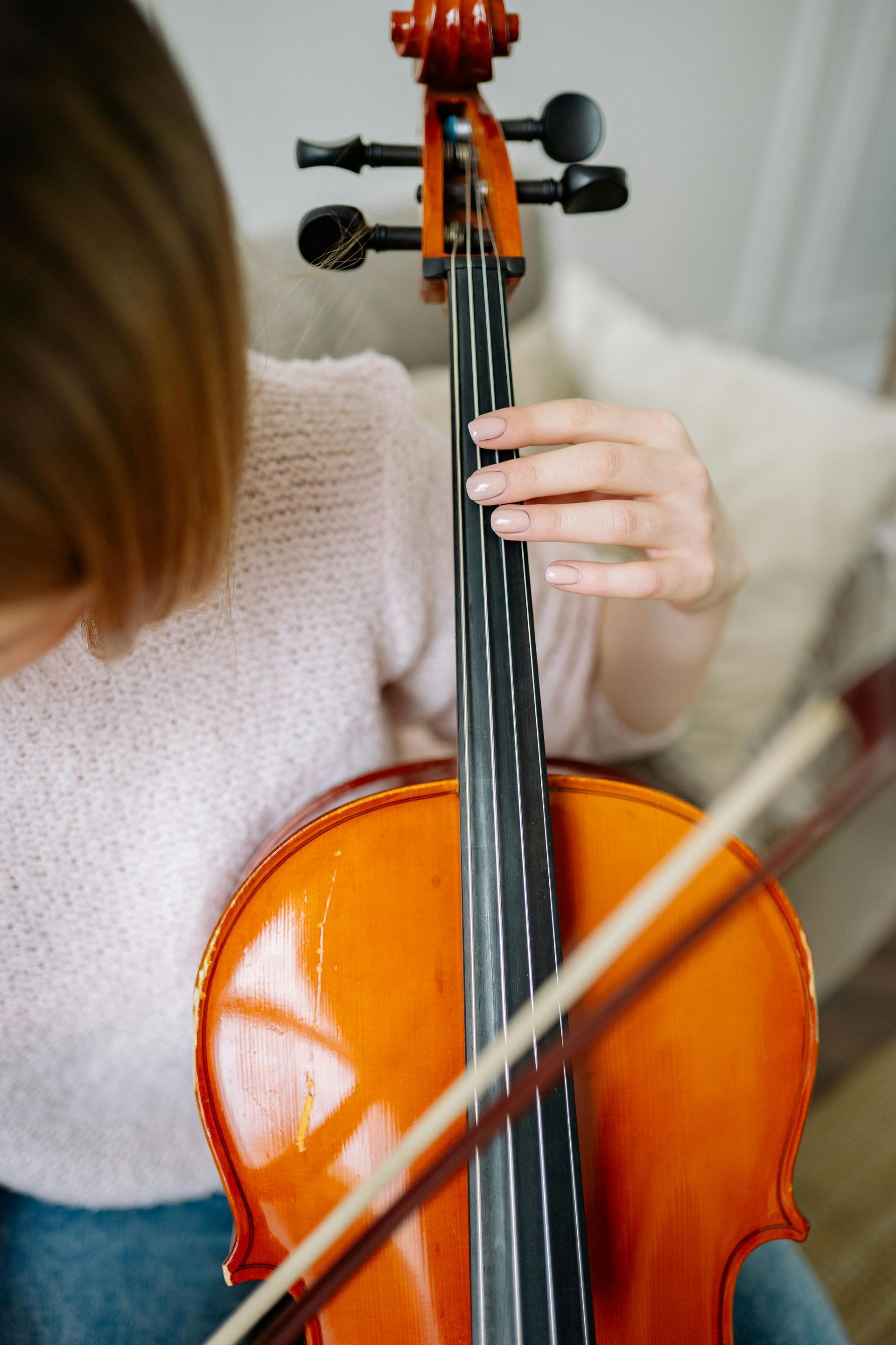 A person in a light pink sweater plays a cello, hand positioned on the fingerboard with a bow drawn across the strings.
