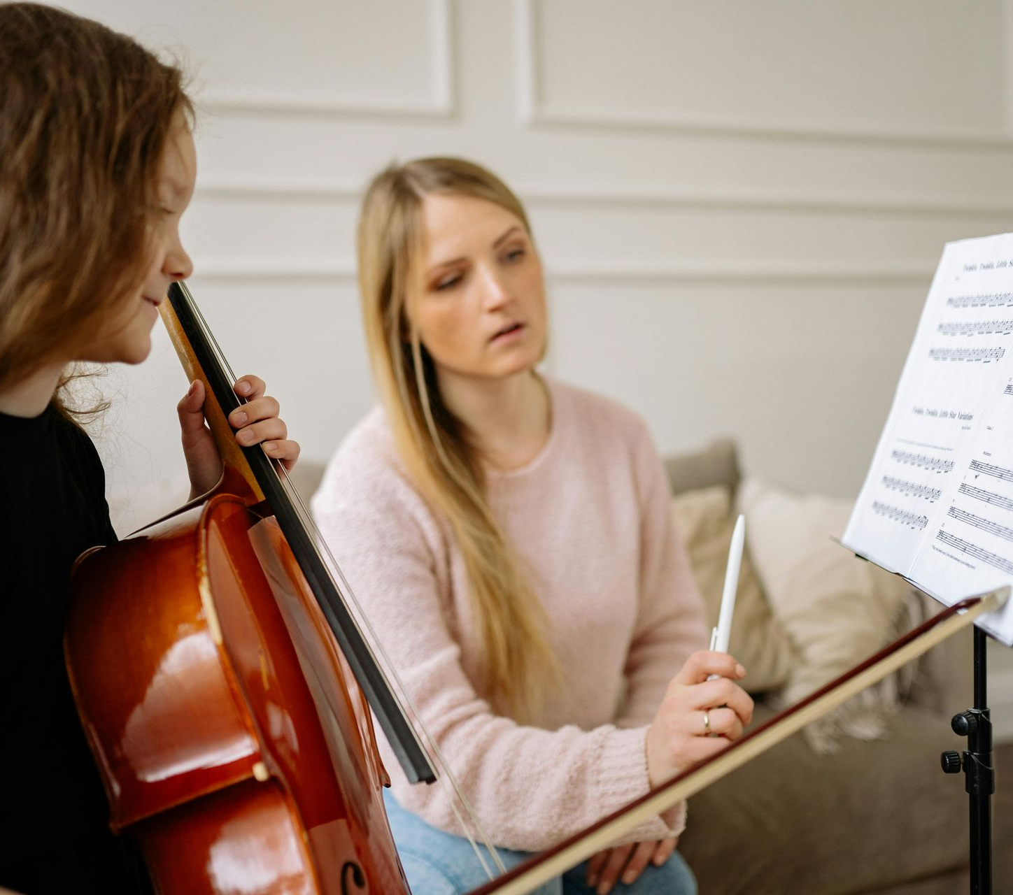 A music teacher points to sheet music on a stand while a student holds a cello during a lesson.