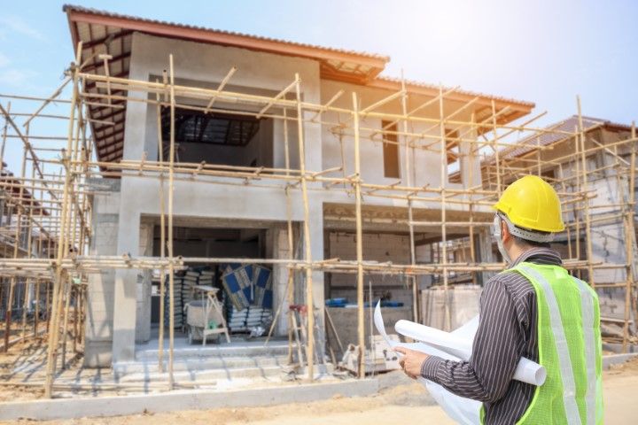 Construction worker in yellow hard hat and vest holding blueprints, observing a house under construction with scaffolding.