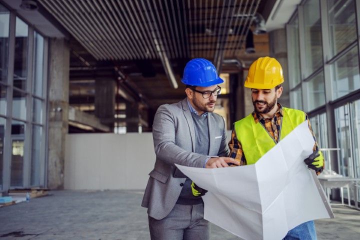 Two men in hard hats reviewing blueprints in a construction site interior. One wears a suit, the other a safety vest.