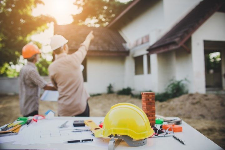 Two construction workers with hard hats discuss plans near a house, tools on a table.