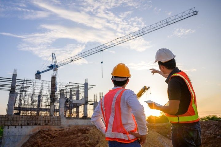 Two construction workers in vests and helmets at a building site, crane overhead, at sunset.