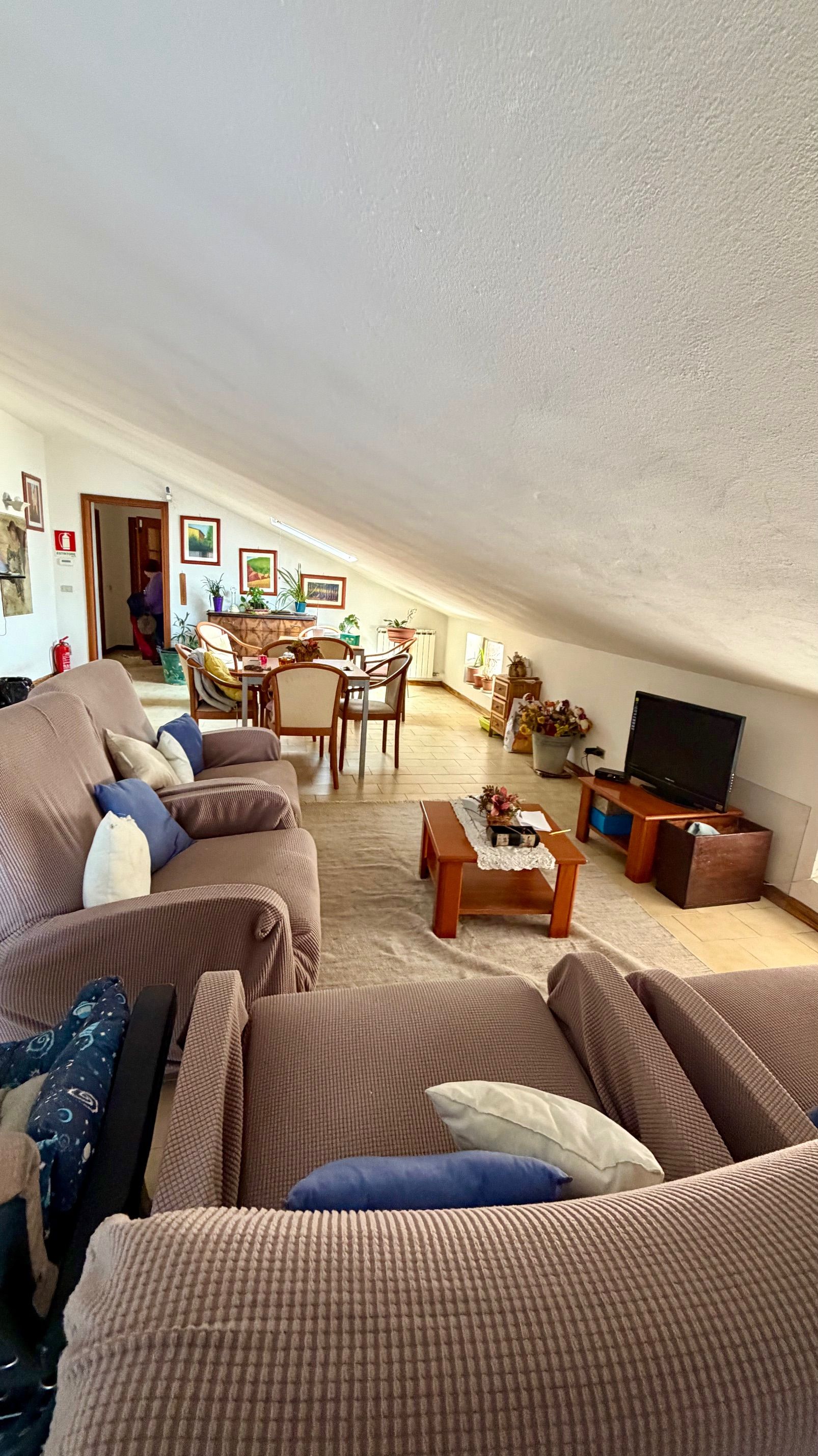 Soggiorno con soffitto inclinato, divani beige, tavolino da caffè, TV e zona pranzo sullo sfondo.