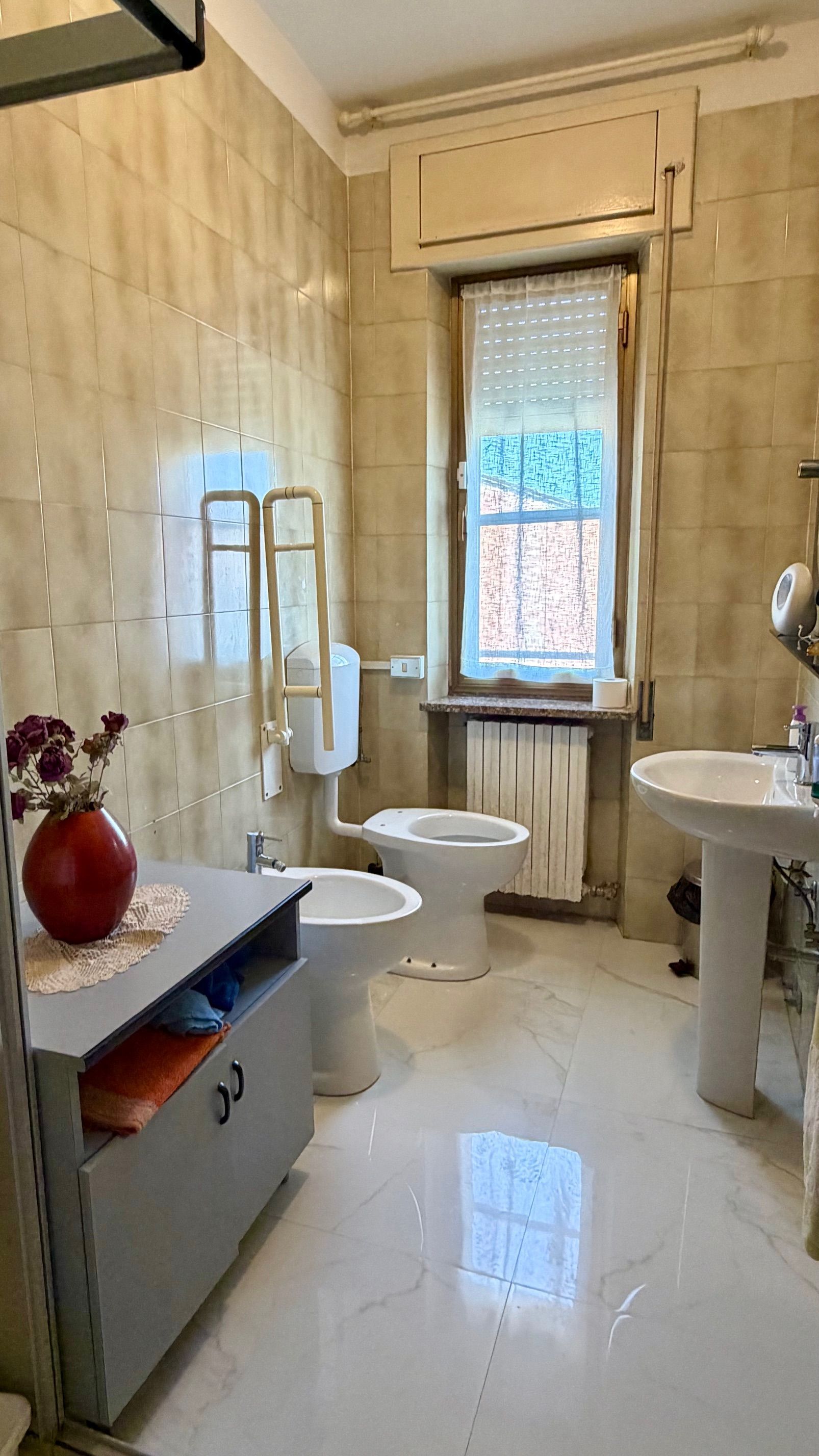 Piccolo bagno piastrellato di beige con finestra, WC, lavandino, bidet e vaso rosso su un mobiletto.
