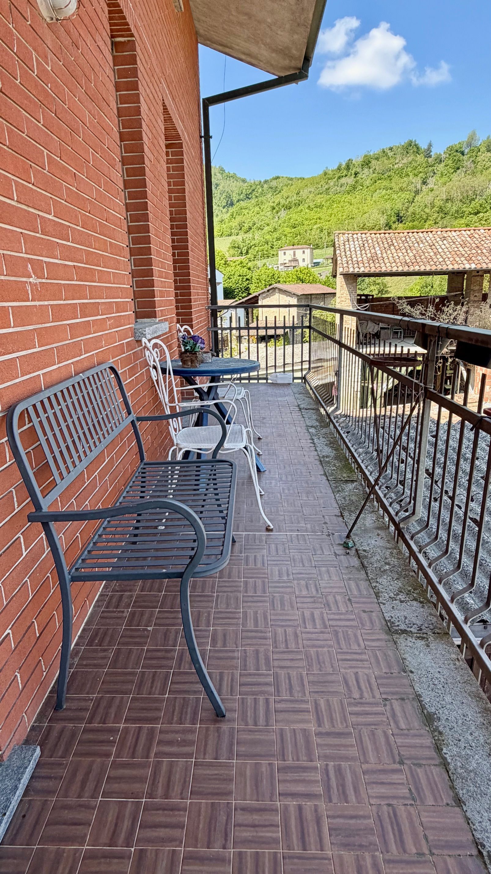 Balcone in mattoni con sedie e ringhiera in metallo nero, affacciato su una verde collina e con area salotto esterna.