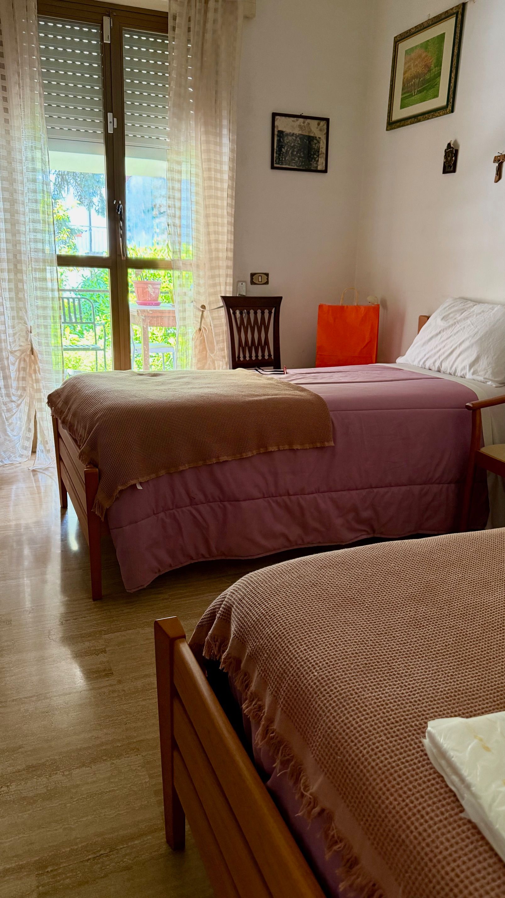 Piccola camera da letto con due letti singoli, sedia in legno e luce naturale che filtra attraverso una porta-finestra sul balcone.
