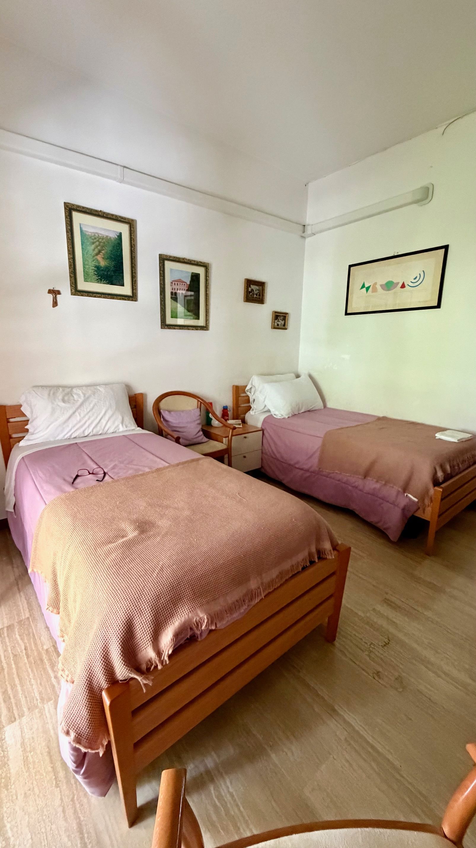 Piccola camera da letto con due letti singoli, lenzuola rosa, quadri alle pareti e una sedia di legno vicino alla finestra.