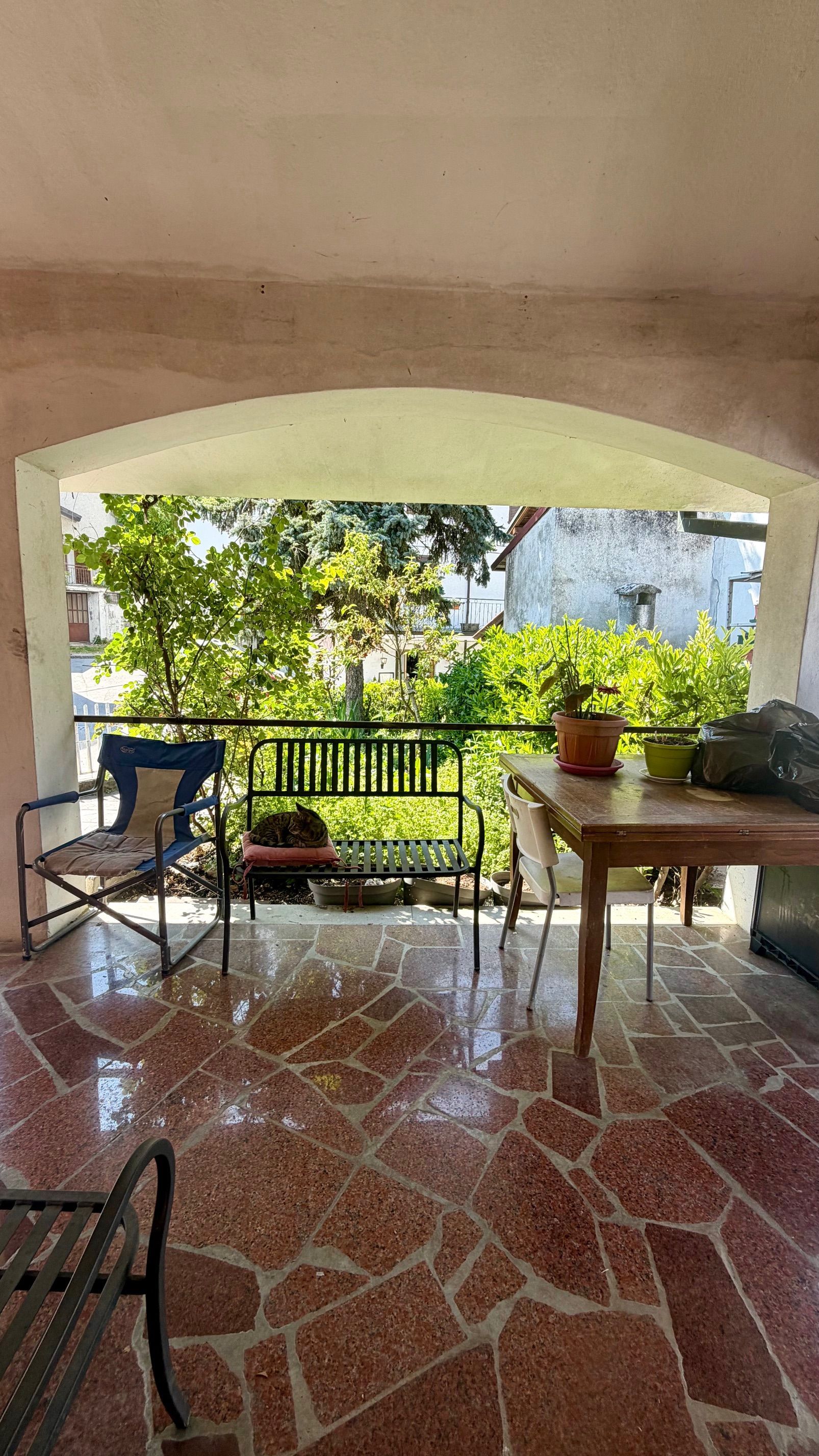 Patio coperto con pavimento in piastrelle rosse, sedie, un tavolo e vista sul giardino oltre una ringhiera.
