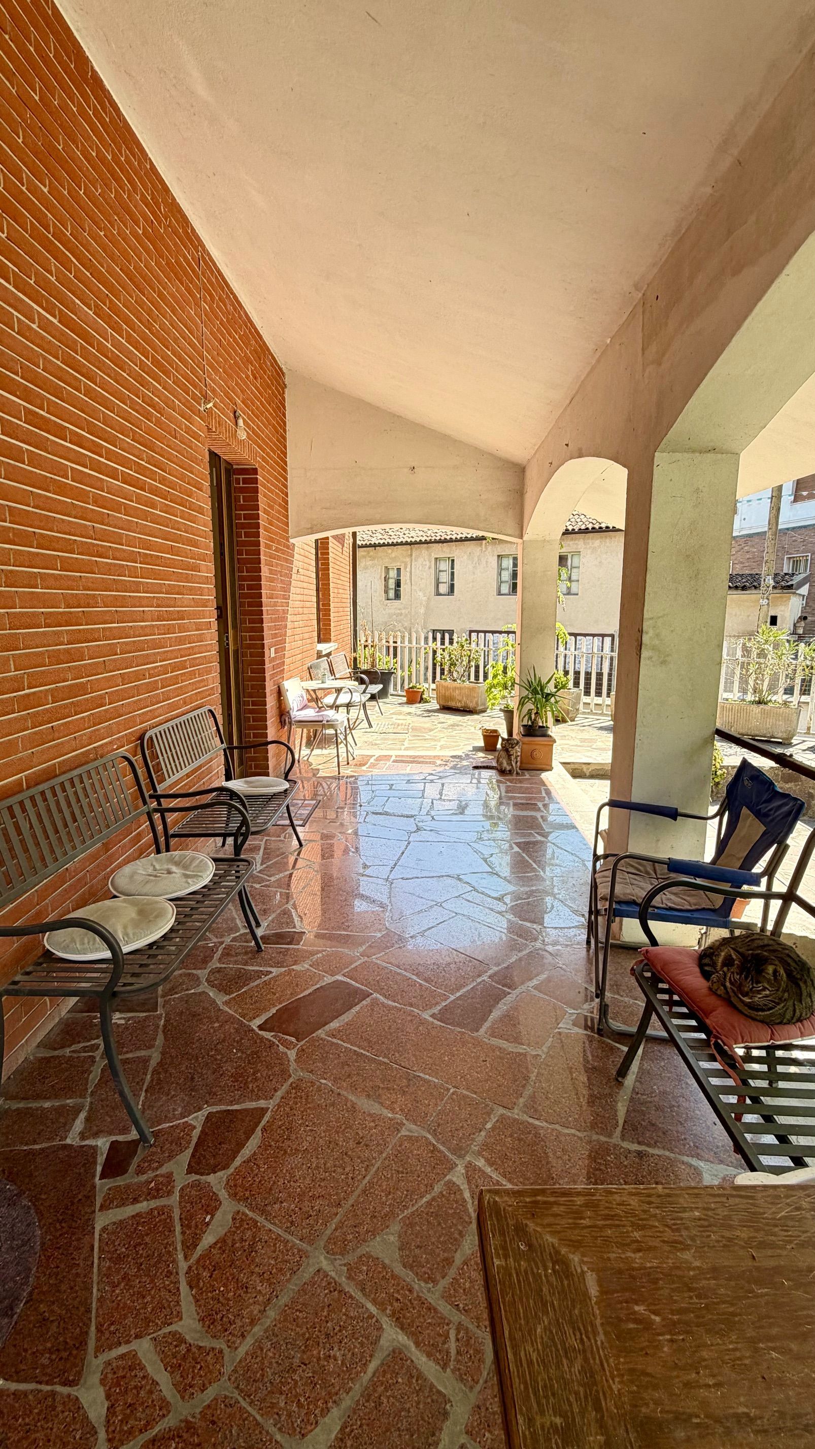 Veranda coperta con pavimento in piastrelle rosse, muro in mattoni e sedie che si affaccia su un cortile soleggiato.