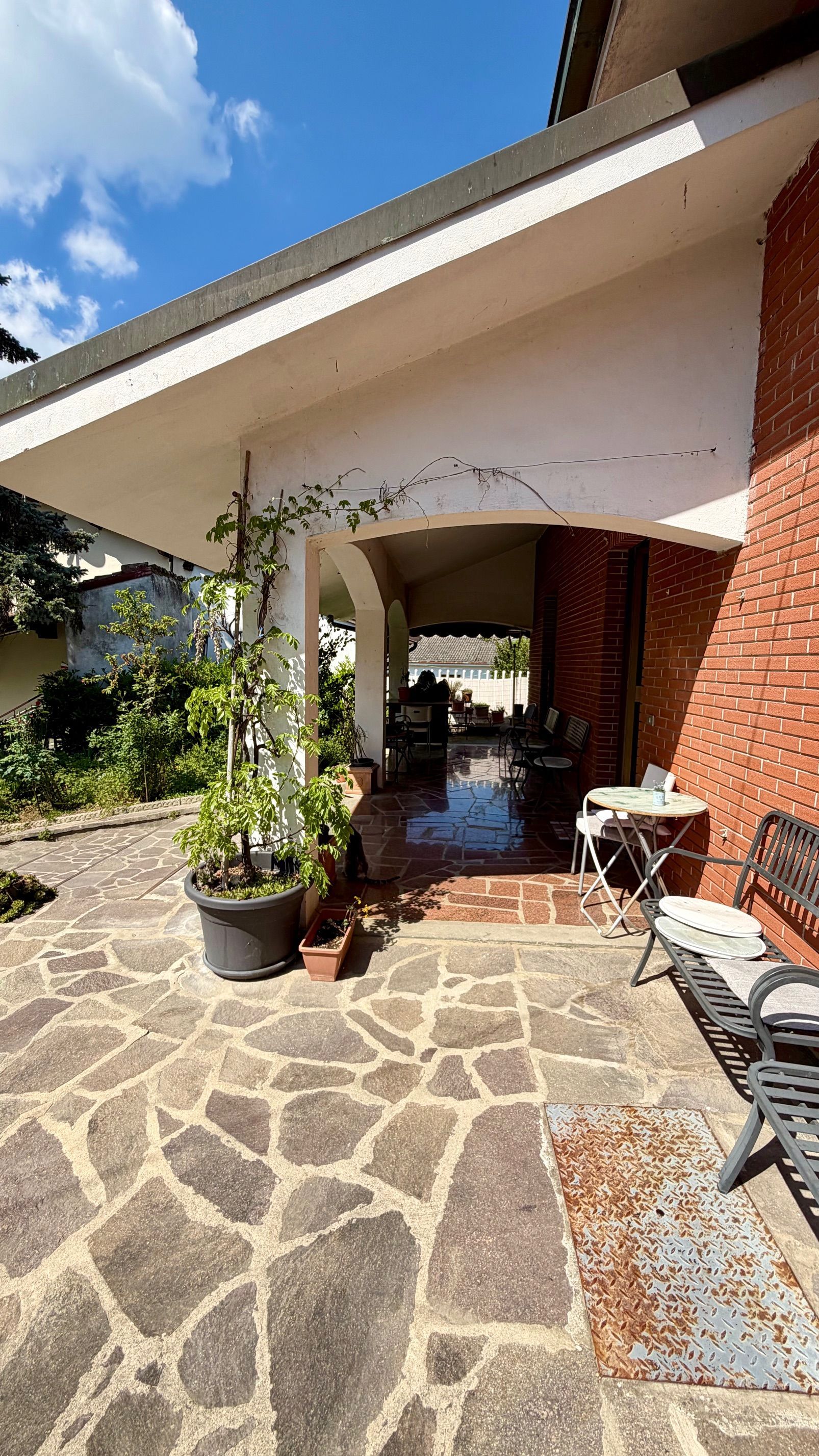 Patio in pietra accanto a una casa in mattoni rossi con piante in vaso, tettoia ad arco e sedie da esterno al sole.