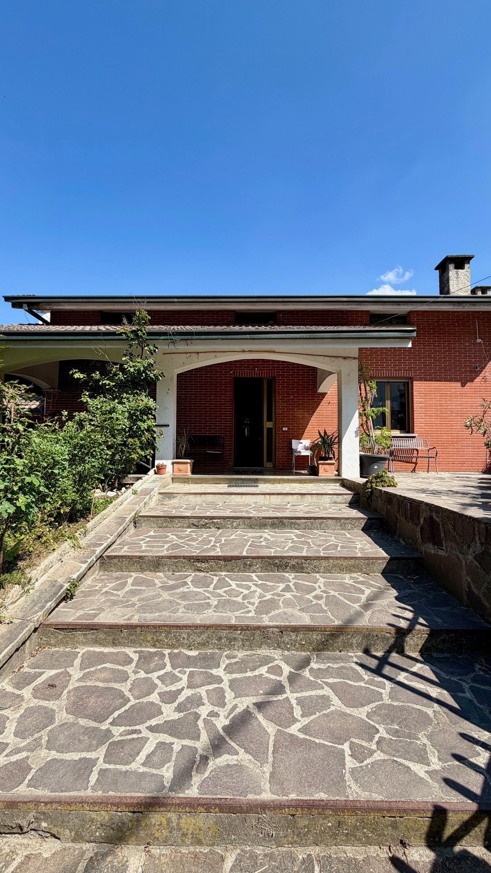 Un sentiero di pietra e una scalinata conducono all'ingresso di una casa in mattoni rossi sotto un cielo azzurro e limpido.