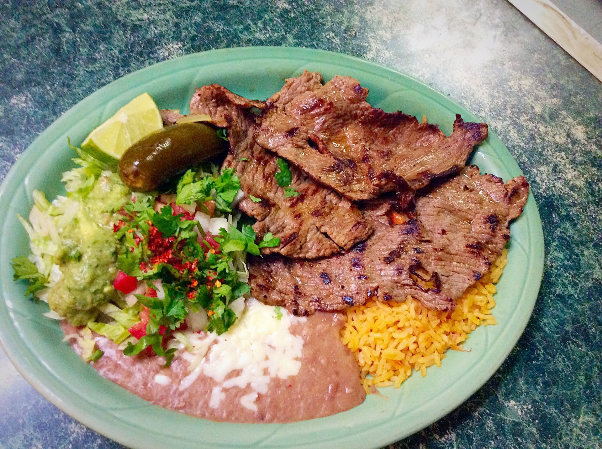 Grilled meat with rice, beans, guacamole, salsa, and a lime wedge on a green plate.