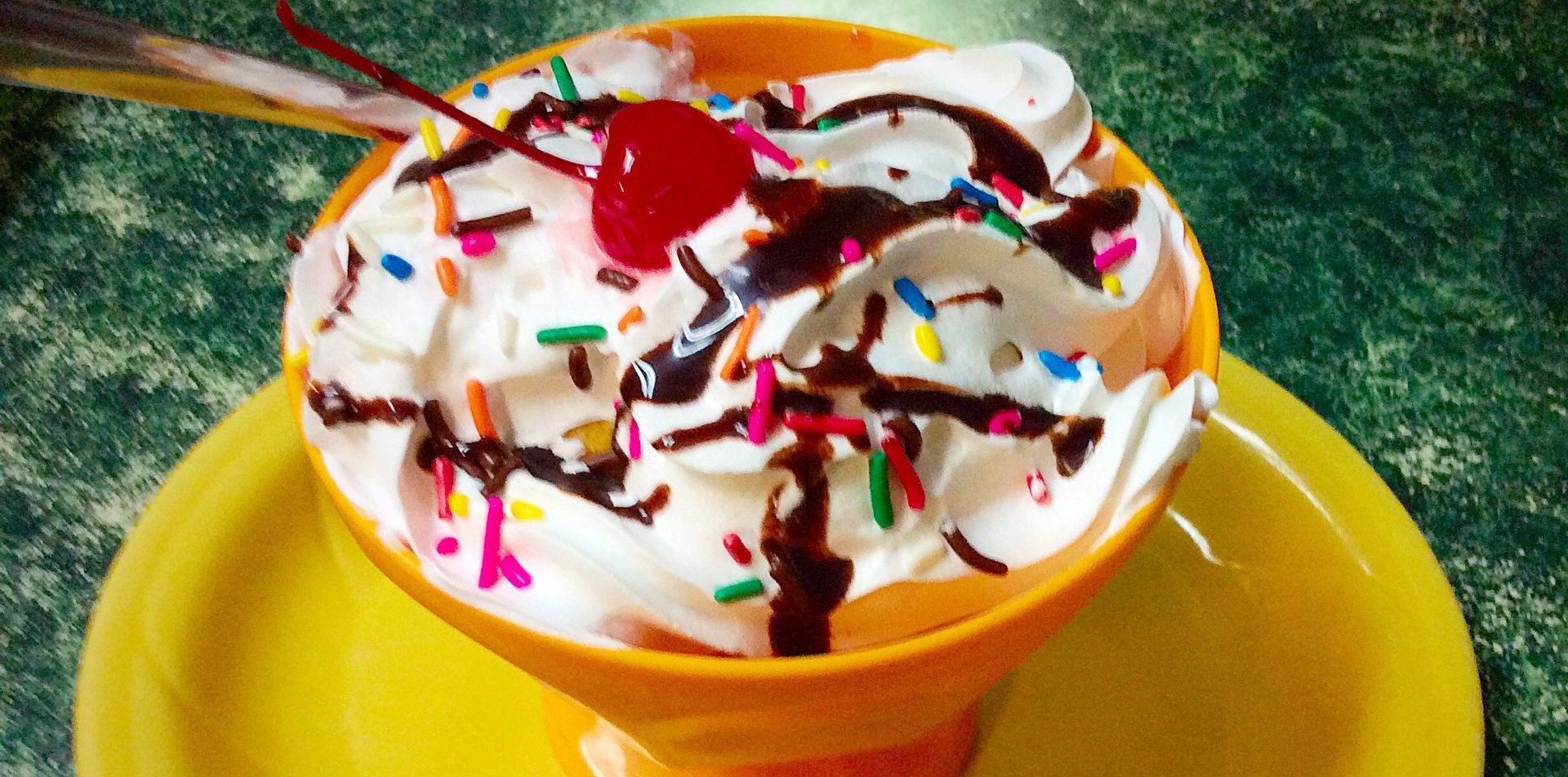 Whipped cream sundae with chocolate syrup, sprinkles, and cherry in orange cup and yellow saucer.