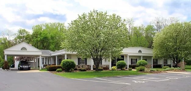 A white building with a carport and a black hearse is parked in the driveway surrounded by trees and green bushes.