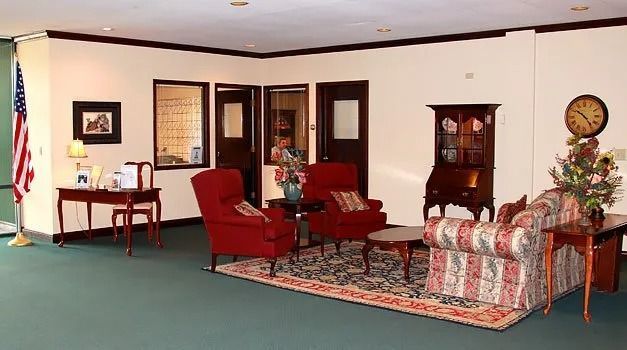 Lobby with red chairs, sofa, antique cabinet, and American flag. Green carpet, cream walls, and brown trim.