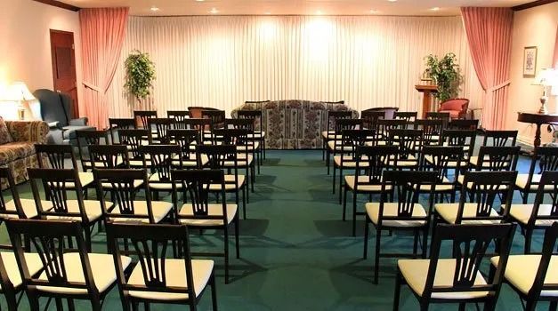 Rows of black chairs face a stage in a formal room, possibly a funeral home. Green carpet, cream curtain backdrop.