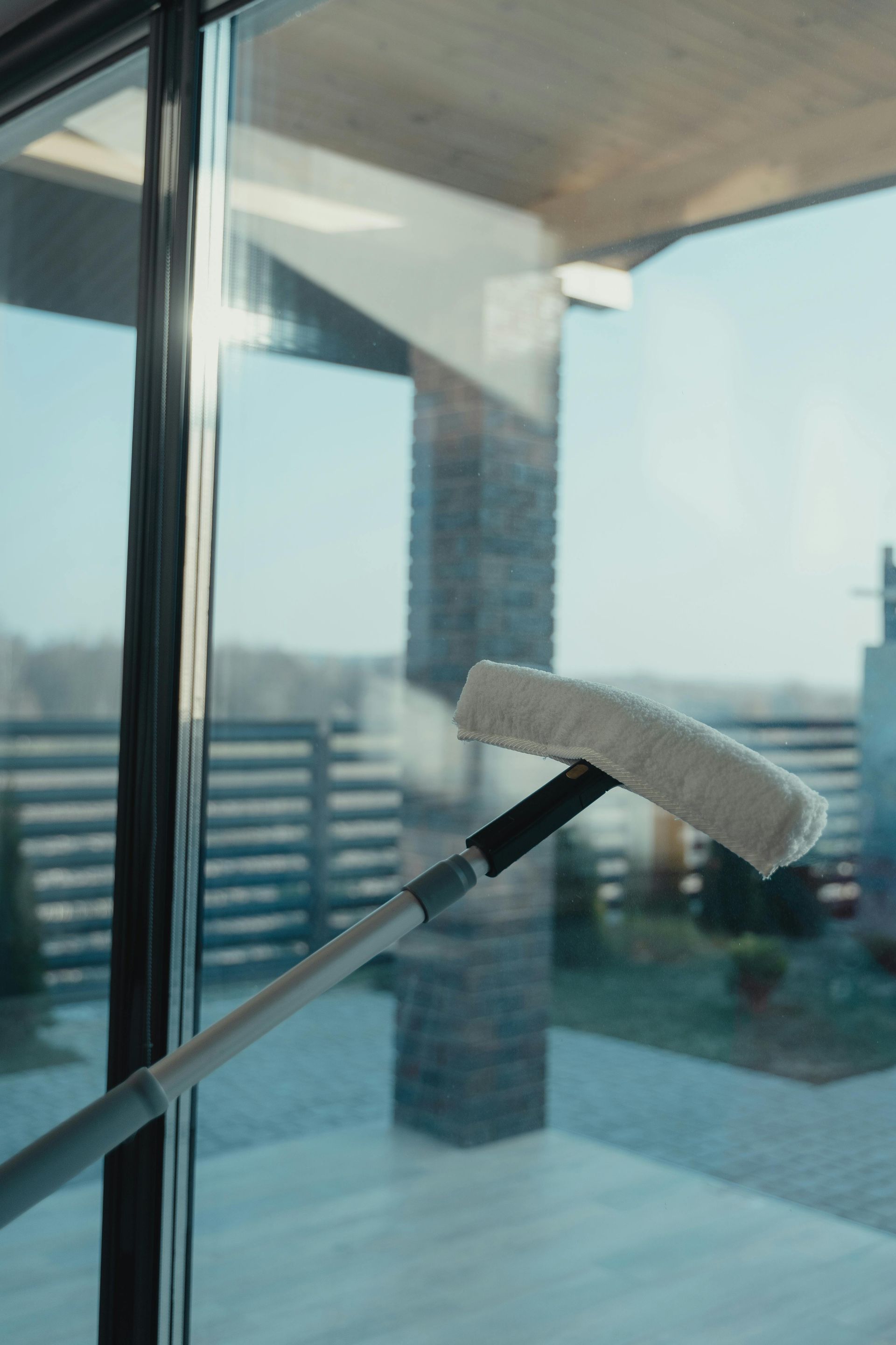 A person cleans a window with a long-handled squeegee, reflecting a house and yard with a brick pillar.