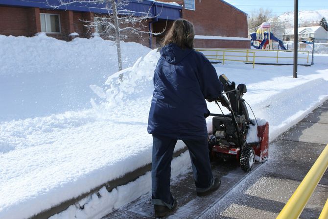 Person shoveling snow beside a curb in a snowy parking lot with a snowblower