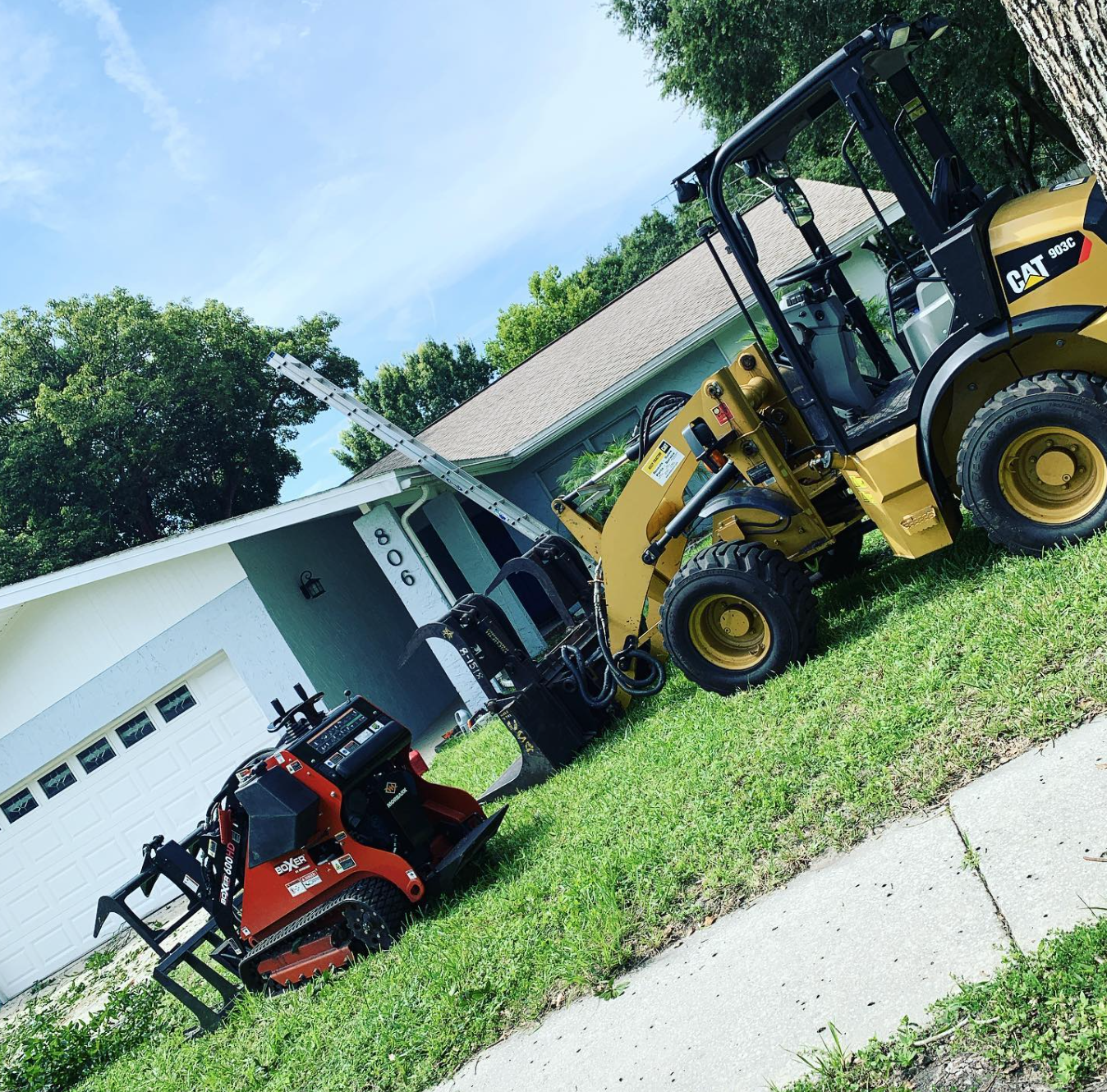 A small yellow Caterpillar loader and red mini-skid steer tractor parked on grass next to a house.
