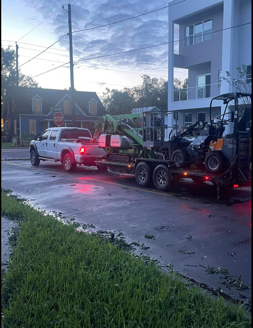 White truck towing a trailer with a green wood chipper and a side-by-side vehicle parked on a street near buildings.