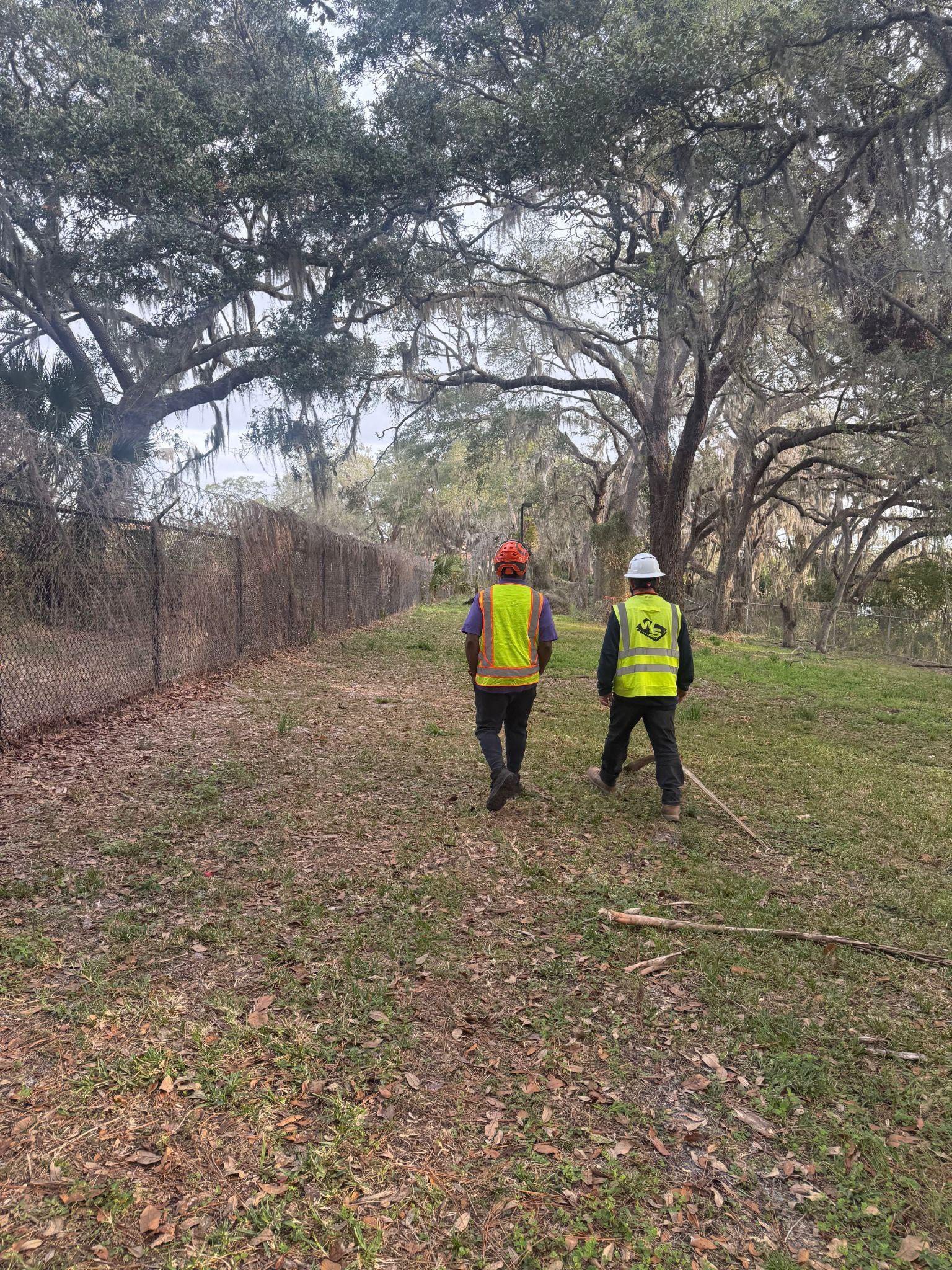 Two workers in safety vests walking on a grassy area, alongside a fence and trees.