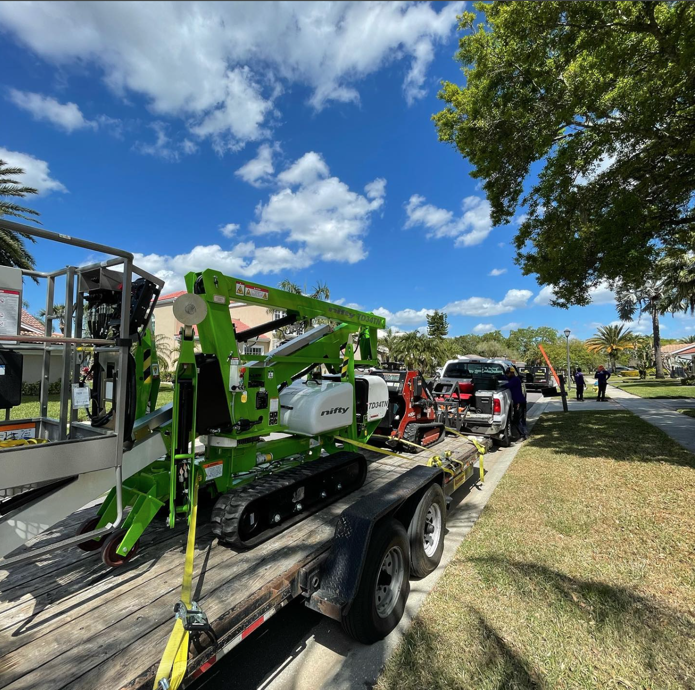 A green and white tracked boom lift on a trailer, next to a white truck on a sunny residential street.