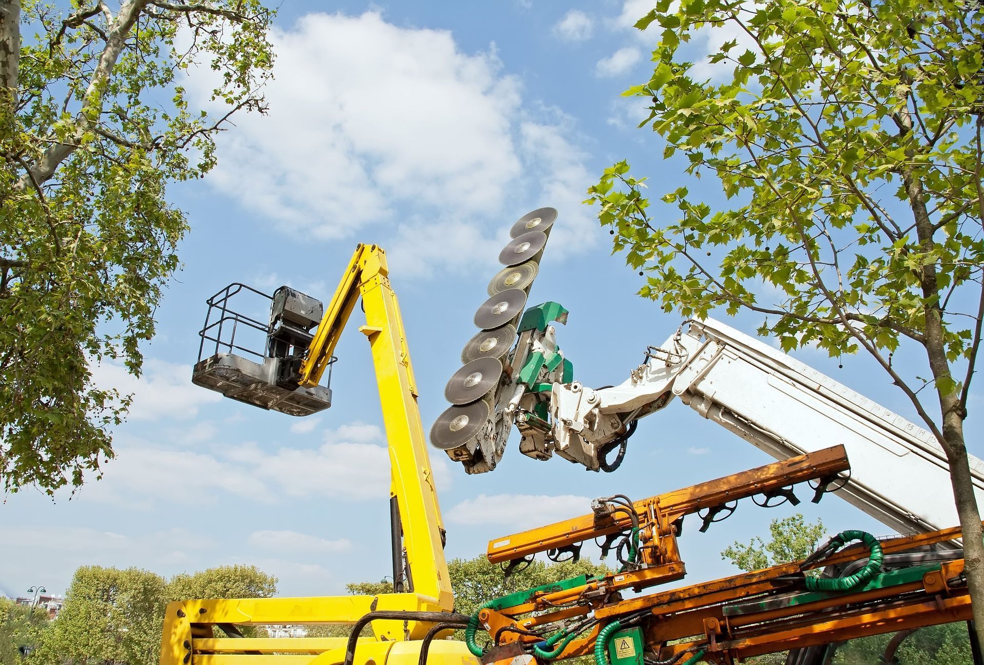 Yellow and white cherry picker with rotating saw blades cutting tree branches against a blue sky.