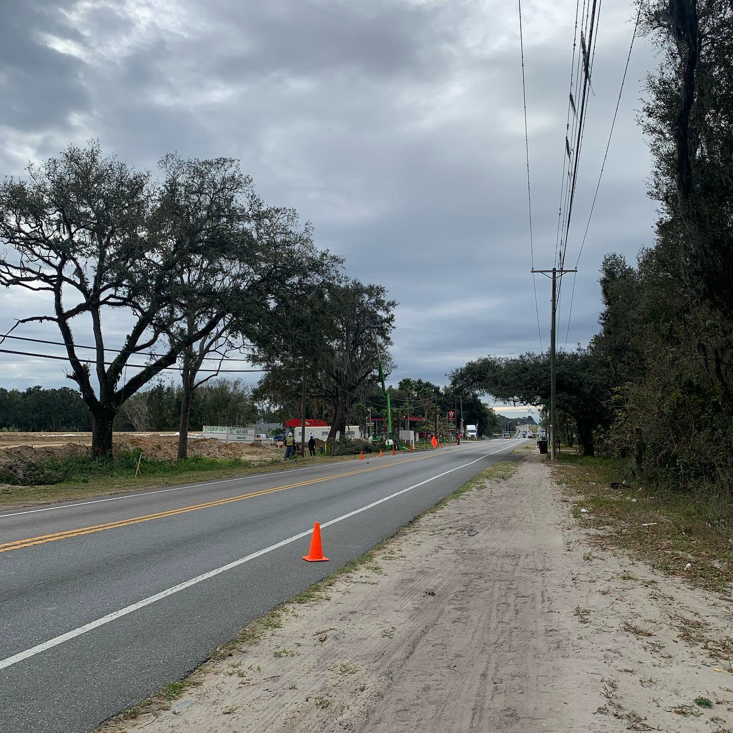 A gray road with a sidewalk beside it, trees, power lines, and an orange traffic cone. Overcast sky.