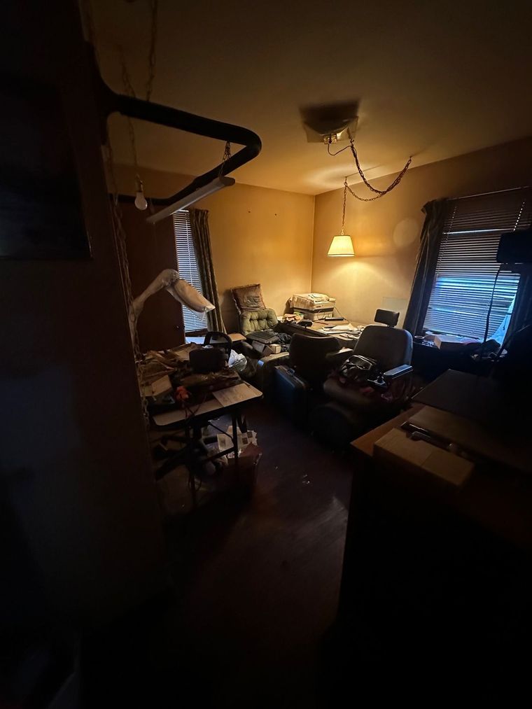 A dark room with a desk , chair , lamp and window.
