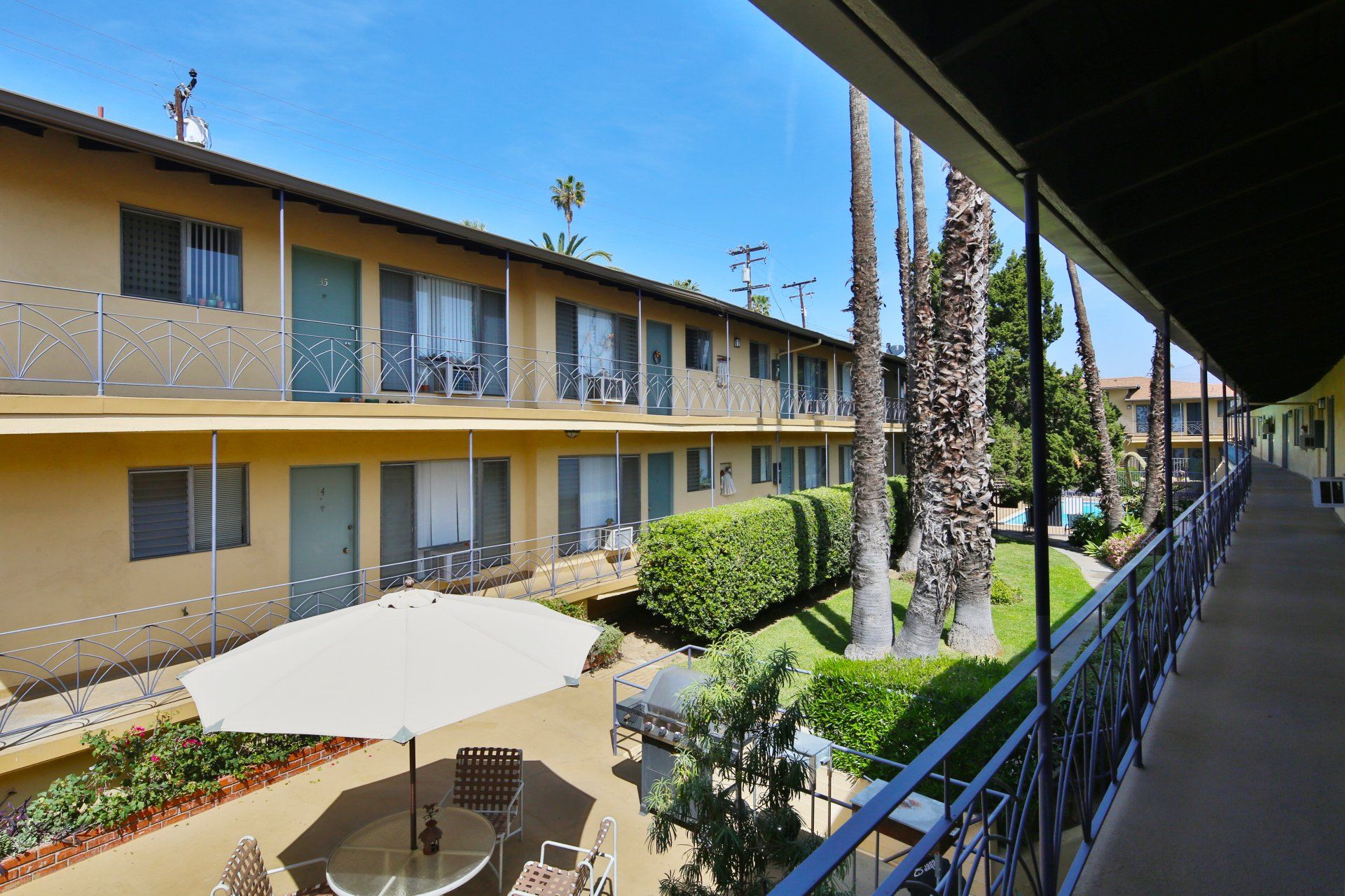 alt="Two-story apartment building with courtyard seating, palm trees, and landscaped walkway."