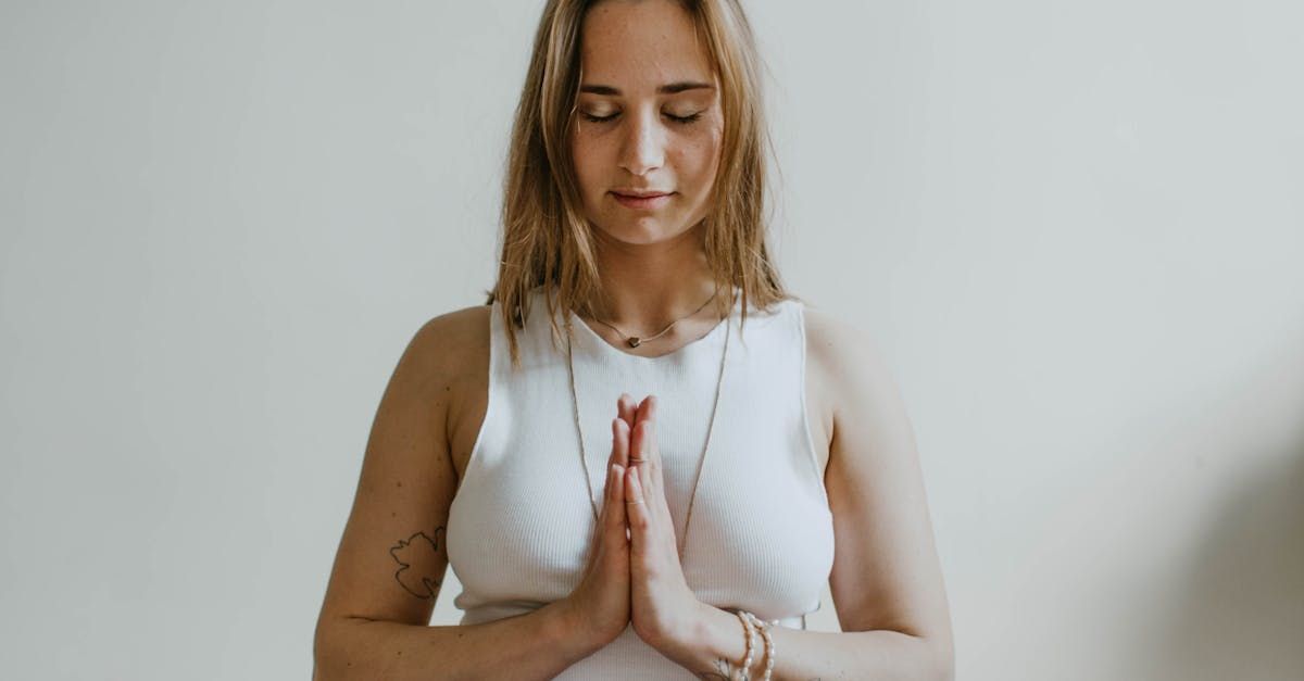 A woman in a white tank top is meditating with her eyes closed.