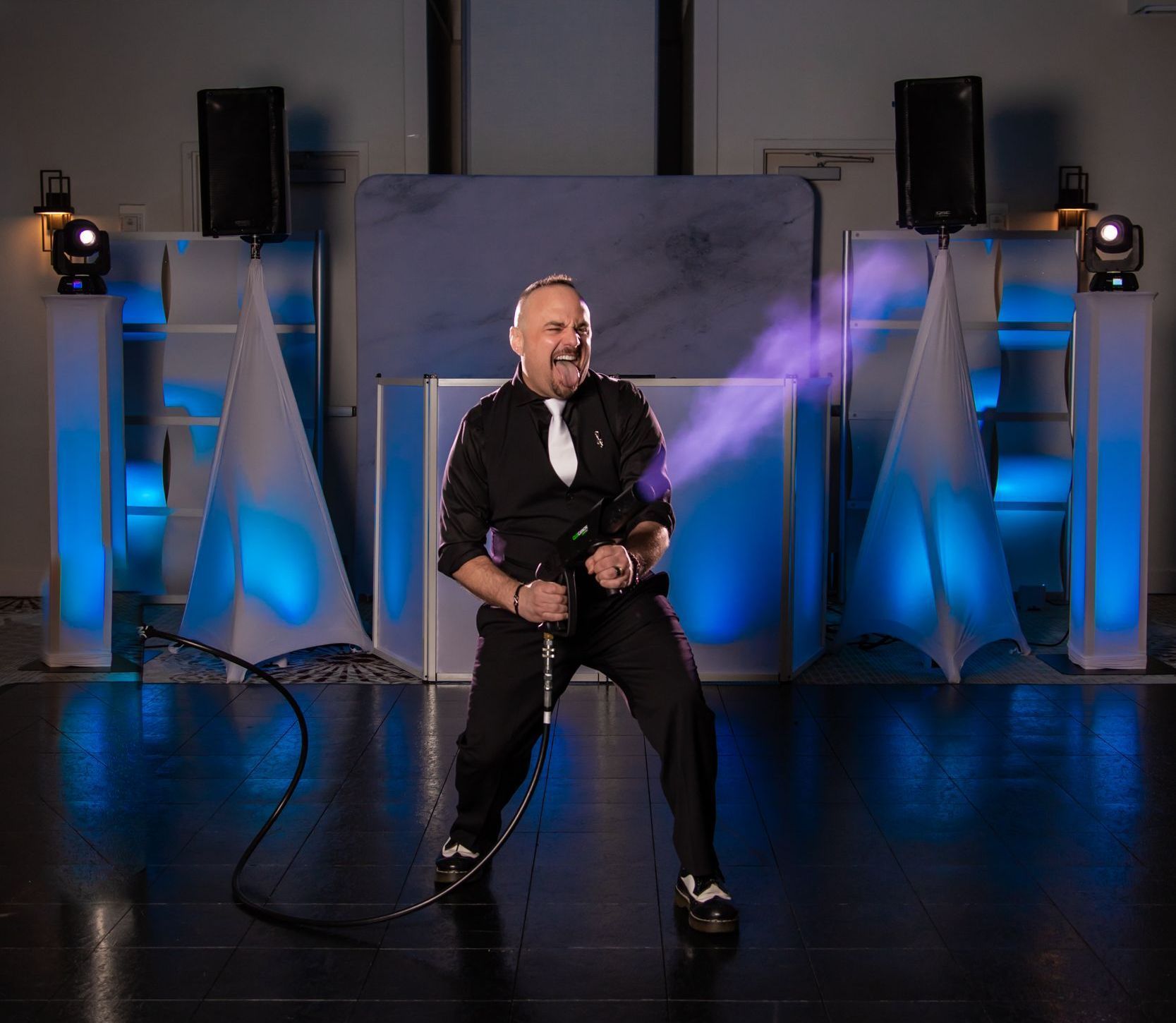 Man holding a smoke gun on a stage, blue and white lights, dark clothing, smiling.
