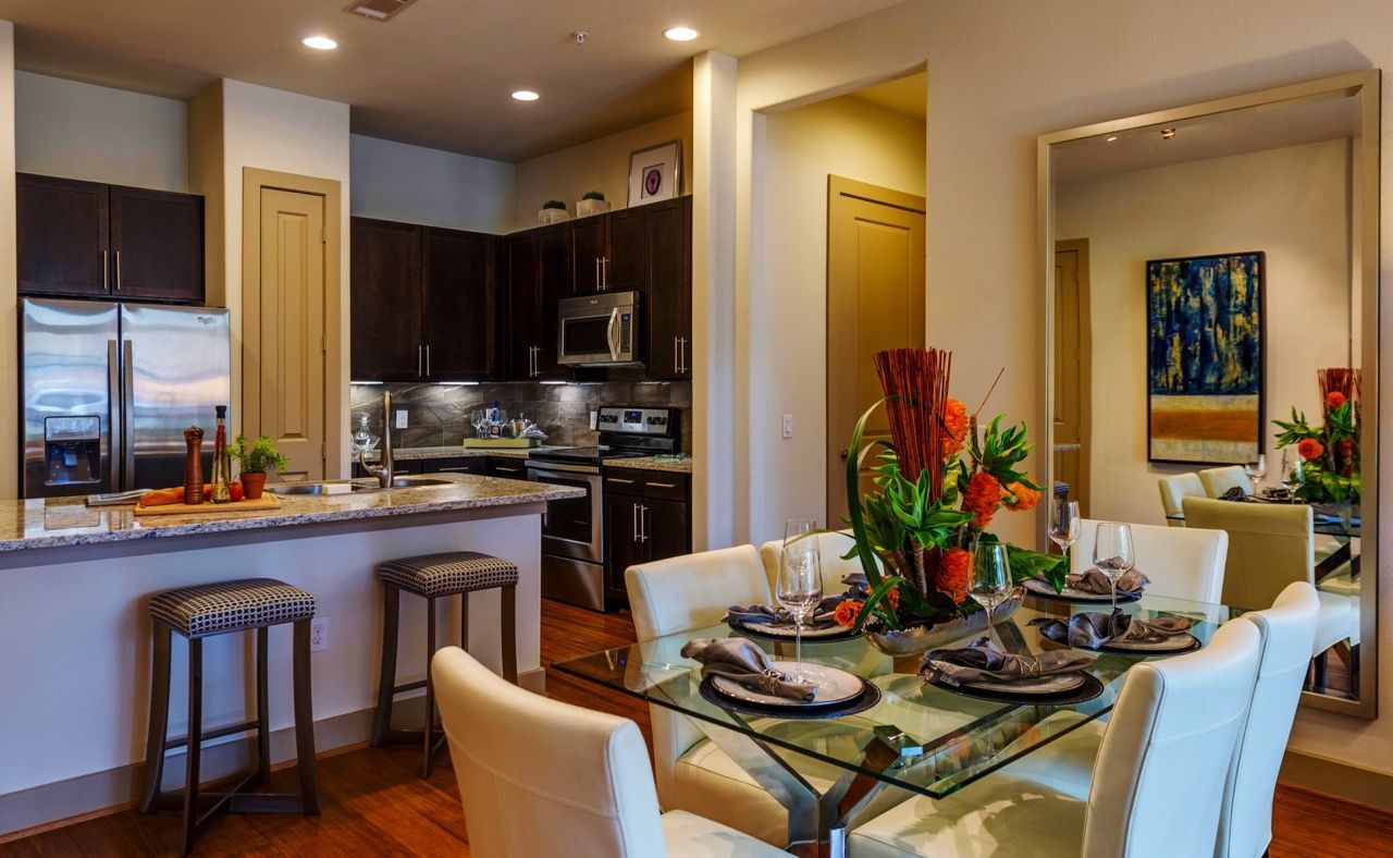 A dining area with a glass table and chairs next to a kitchen with dark cabinets, stainless appliances, and a breakfast bar.