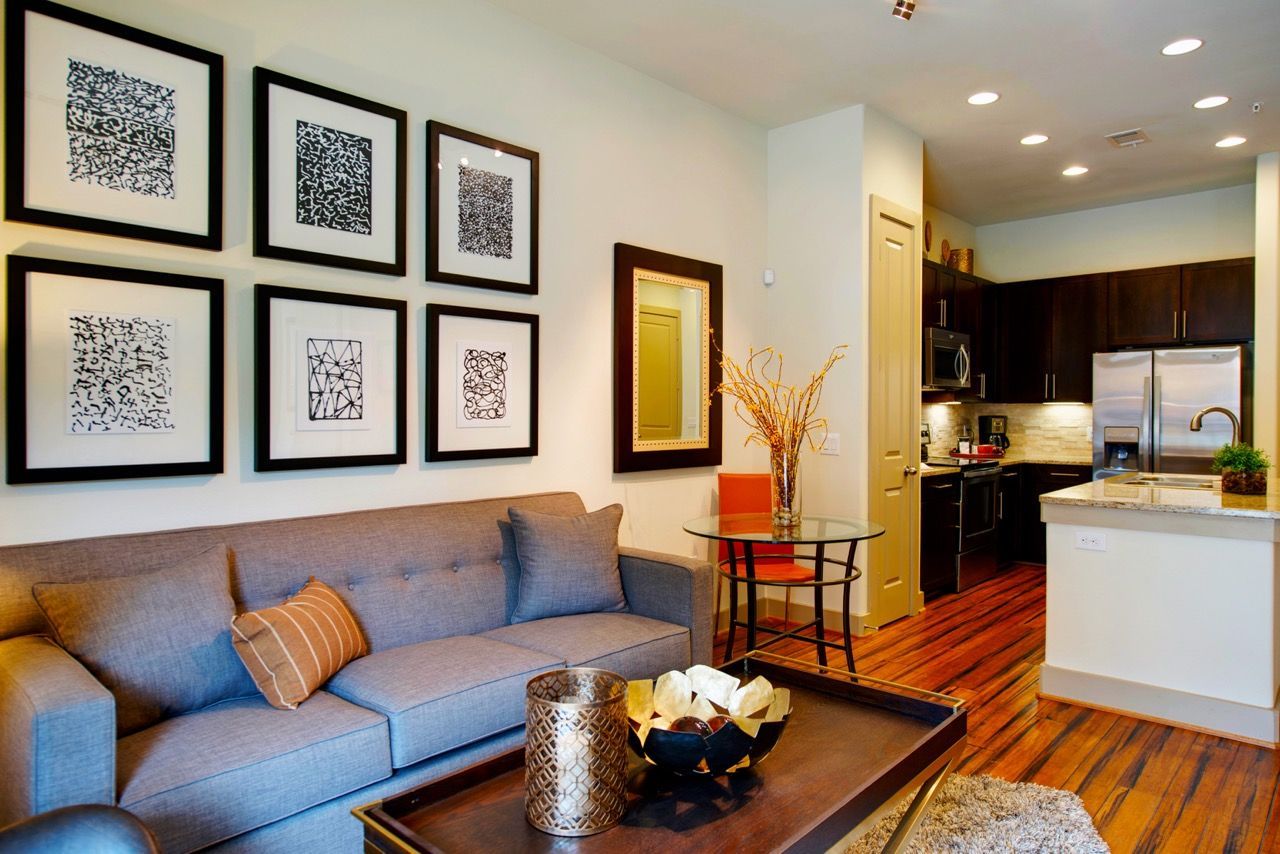 Open-concept living room with a gray sofa, six framed art pieces on the wall, and a round glass dining table.