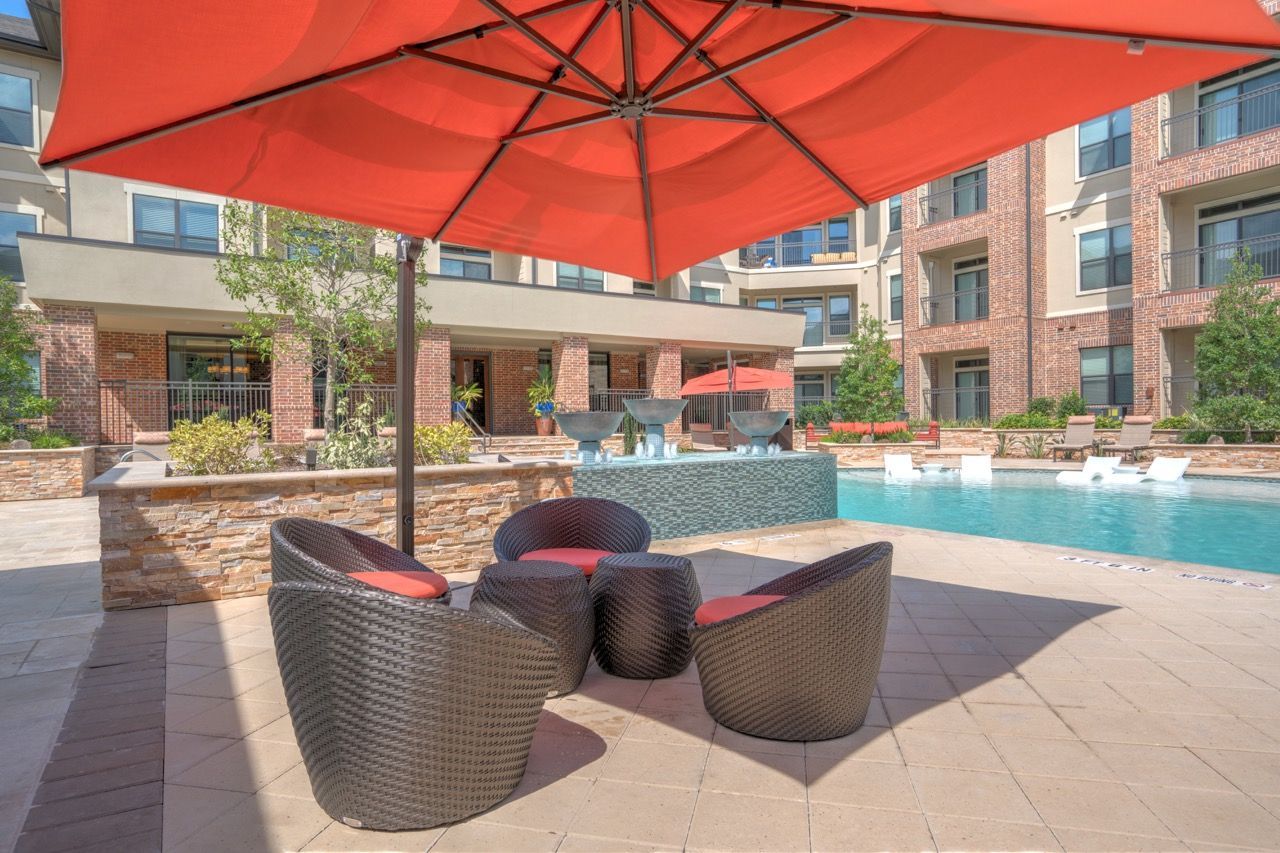 An orange patio umbrella shades brown wicker chairs and a table on a stone pool deck near an apartment complex.
