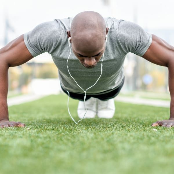 A person wearing a grey t-shirt and wired headphones does a push-up on a green outdoor field.