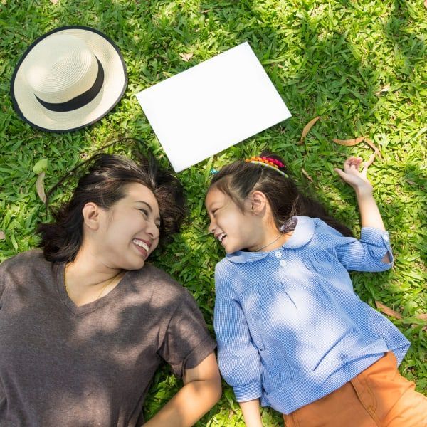 A woman and child lie on green grass, smiling at each other with a blank white paper and a straw hat nearby.